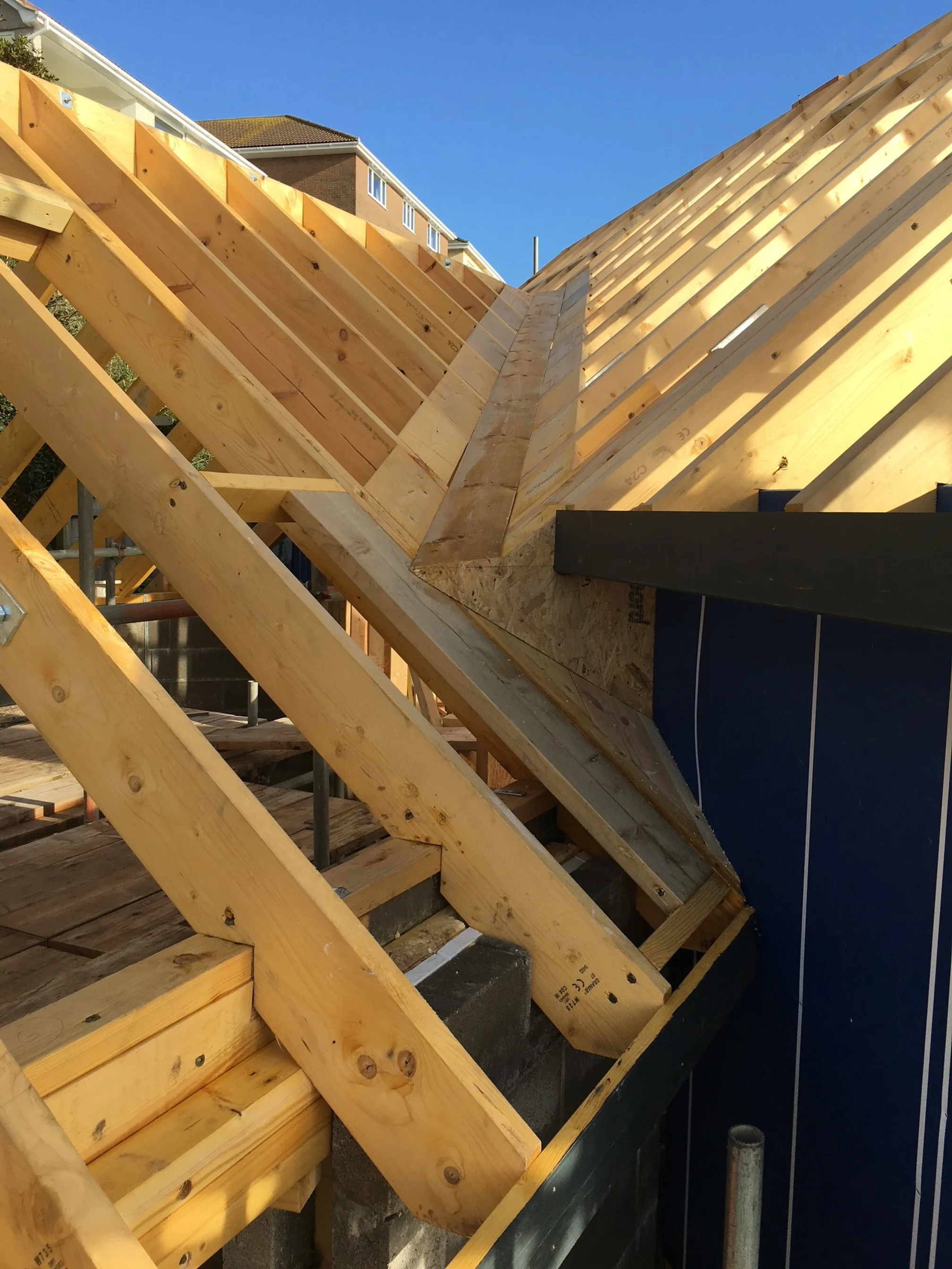Close-up of a roof frame under construction with wooden beams and plywood sheathing, part of a building with neighboring houses and a clear blue sky in the background.