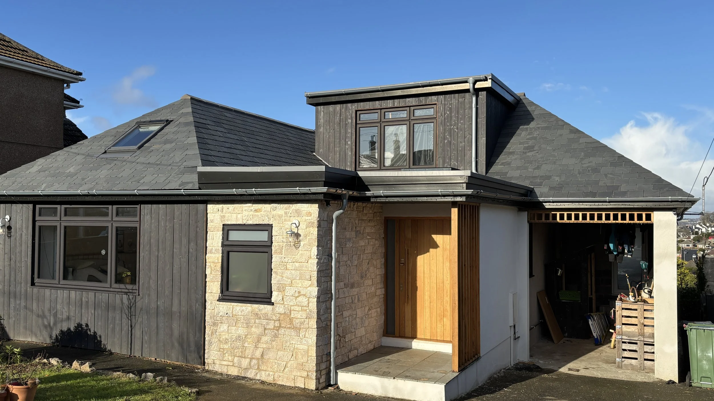 Front view of a modern house with a combination of brick, wood, and white exterior walls, featuring a wooden front door, grey roof, and a garage or carport area with tools and equipment.