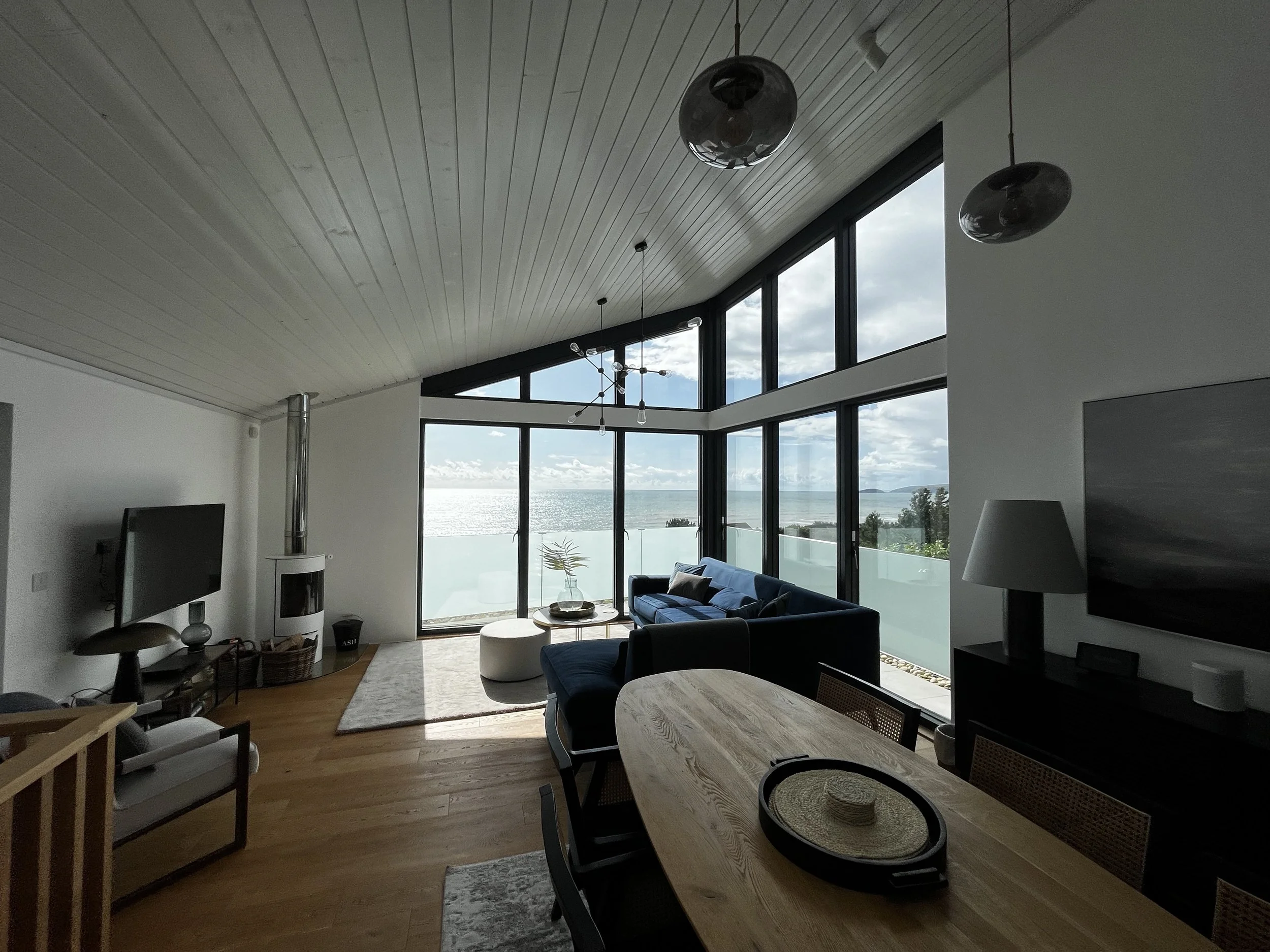 Living room with large floor-to-ceiling windows overlooking an ocean view, with a blue sofa, a white round ottoman, a small coffee table, and a wood dining table with chairs. Modern light fixtures hanging from the ceiling, and a flat-screen TV on a black stand.