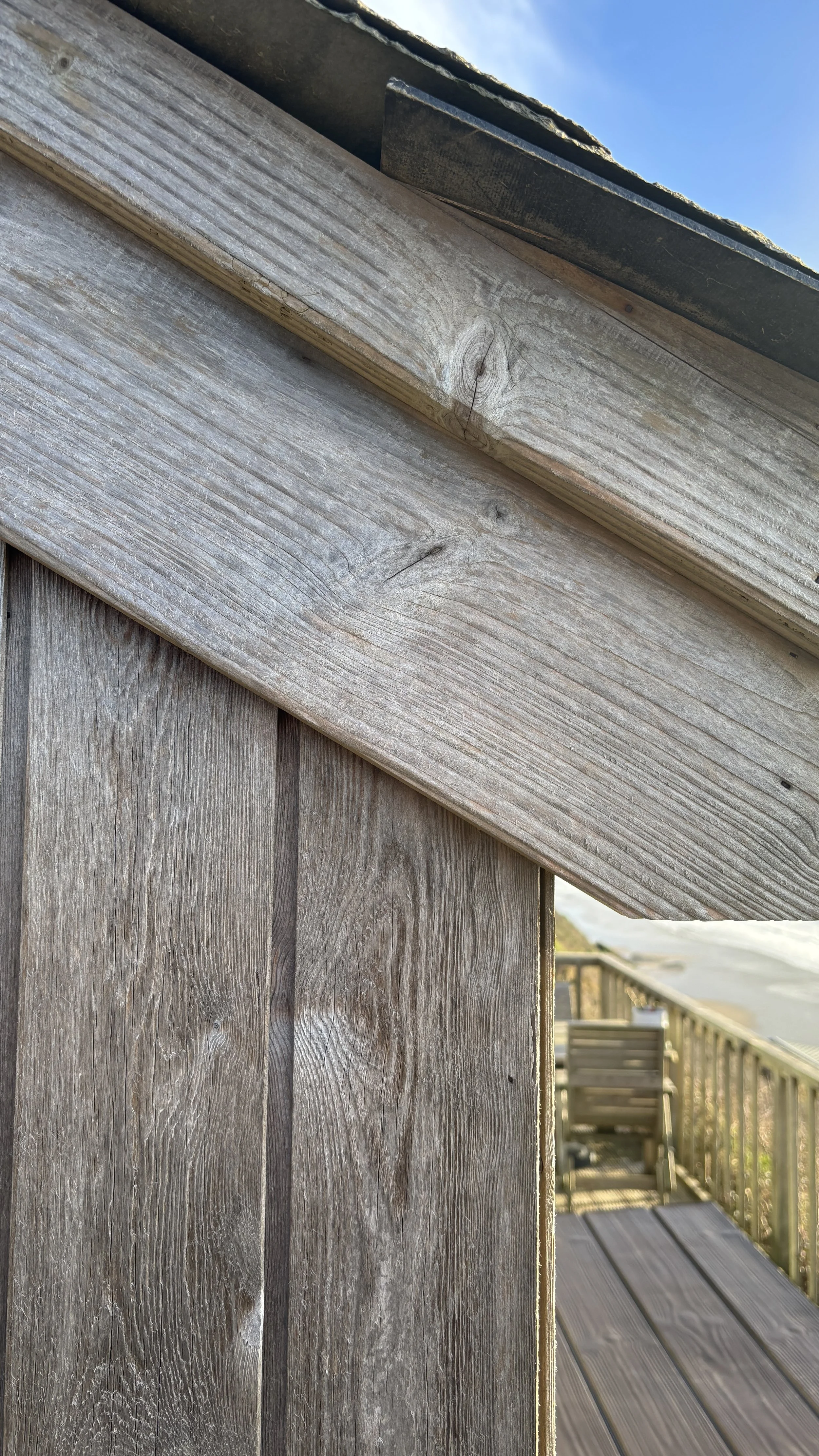 Close-up of wooden siding and corner of a deck with a chair on a balcony in the background, under a partly cloudy sky.