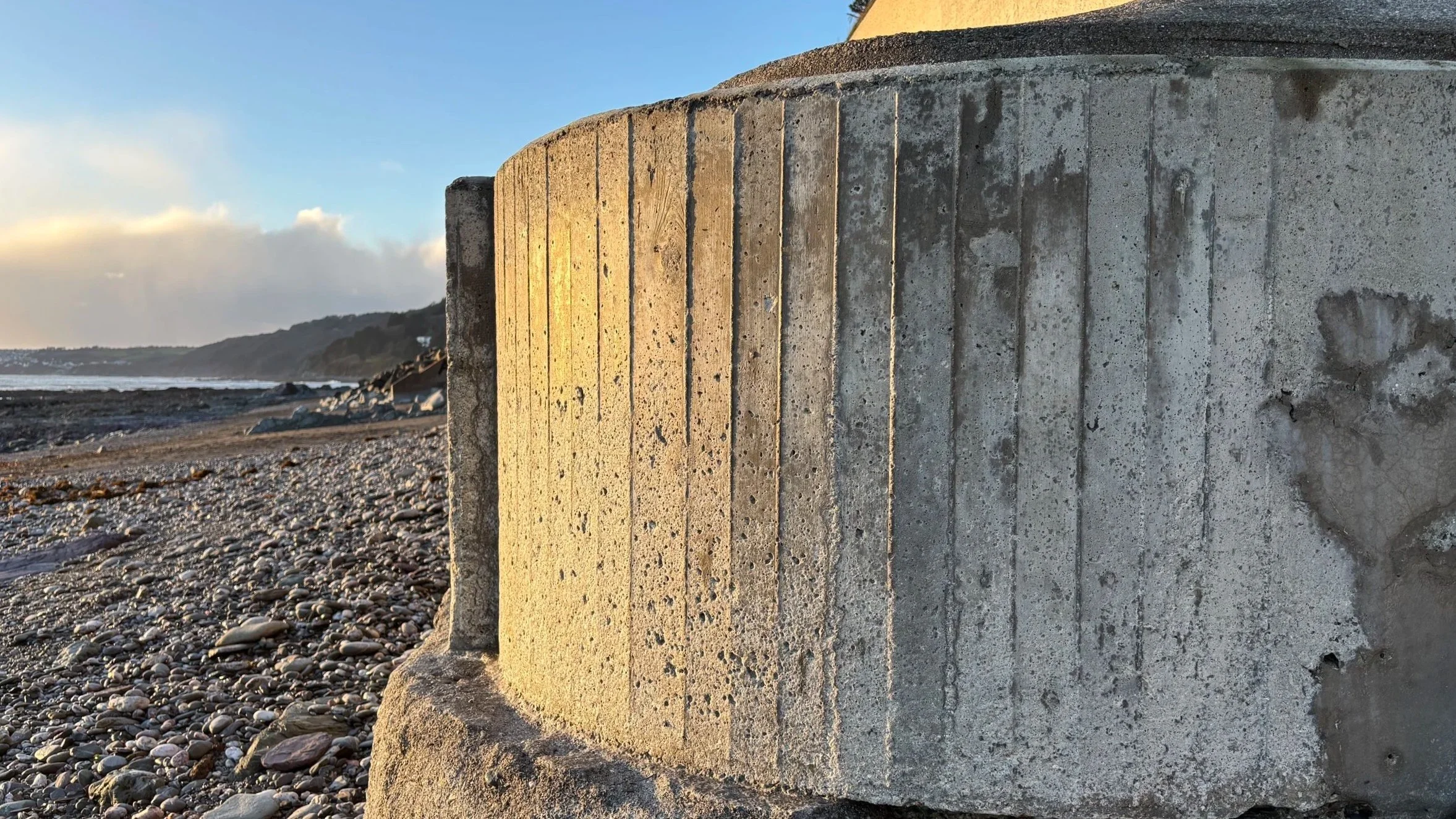 Close-up of a cylindrical concrete structure on a pebble beach during sunset, with the ocean and distant hills in the background.