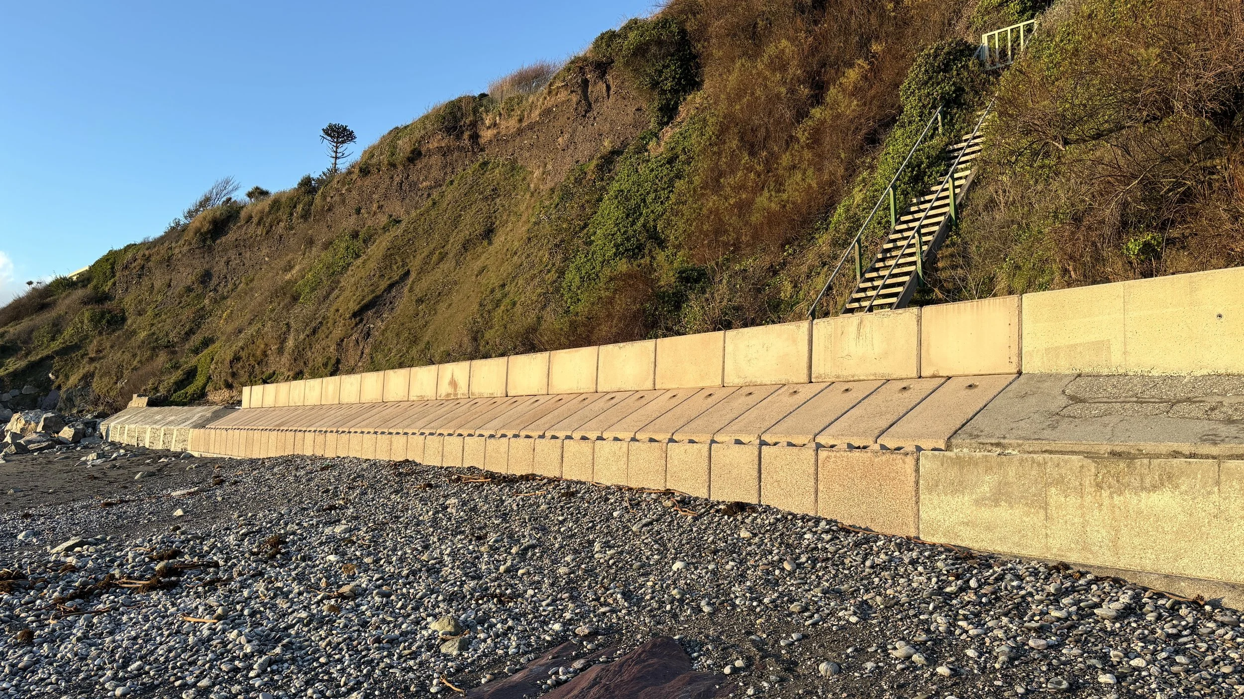 A rocky beach with a concrete seawall along the shoreline, a hillside covered with bushes and trees in the background, and staircases leading up the hillside, with clear blue sky overhead.