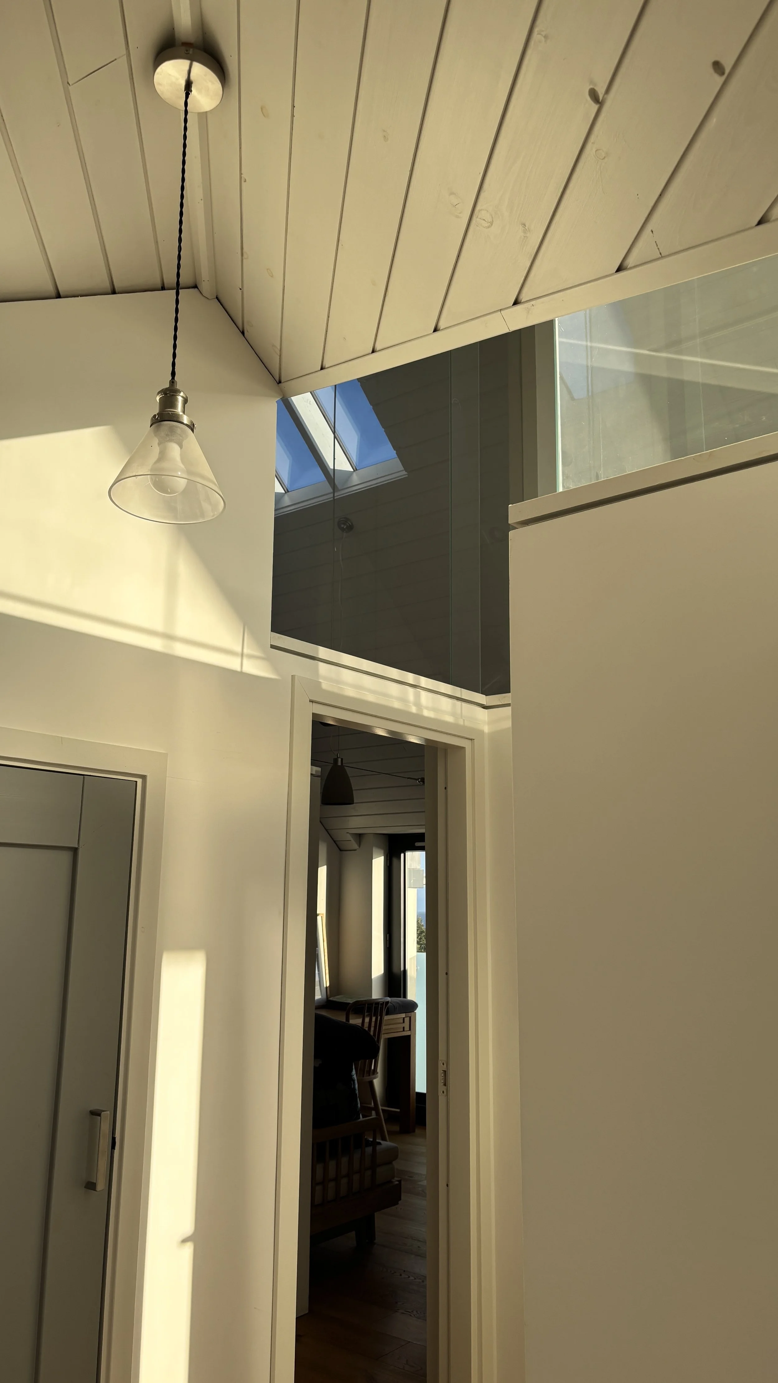 Interior view of a modern house with white walls, wooden ceiling, and a glass wall/rail. Sunlight streams in, casting shadows on the wall, and part of a room with furniture is visible through an open doorway.