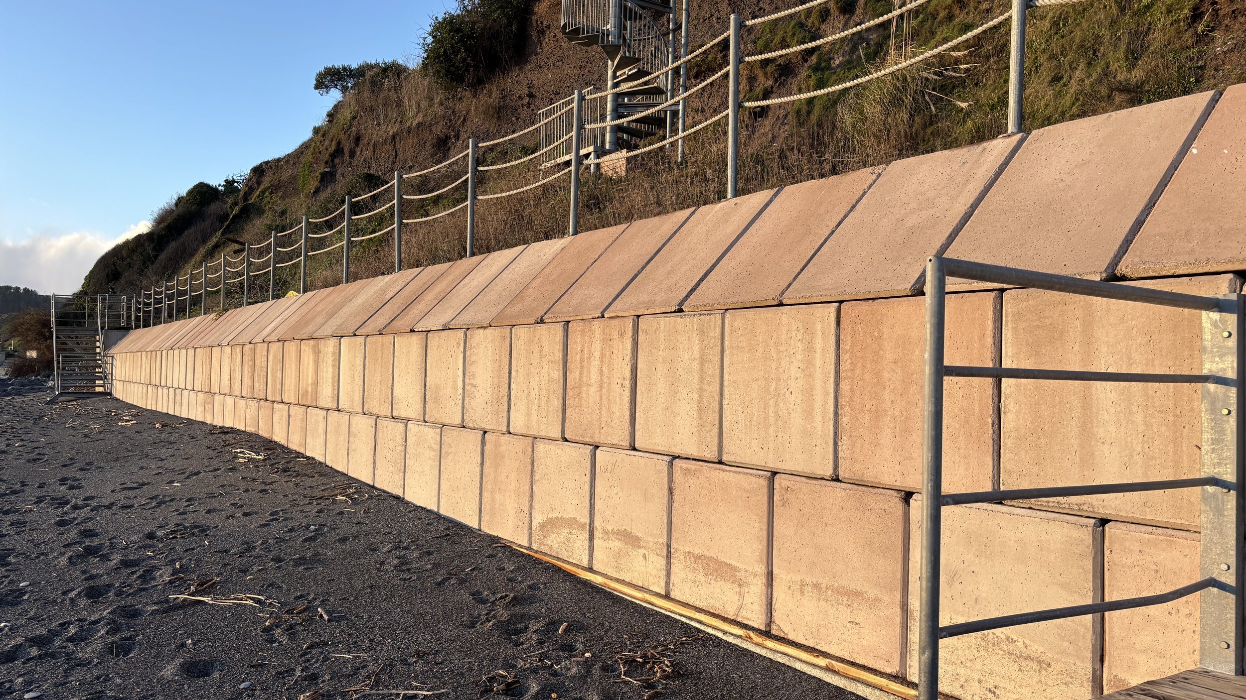 A constructed terraced retaining wall with large concrete blocks separating a hill from a walkable area. Metal fencing and stairs are at the top of the hill, with sparse vegetation and a clear sky in the background.