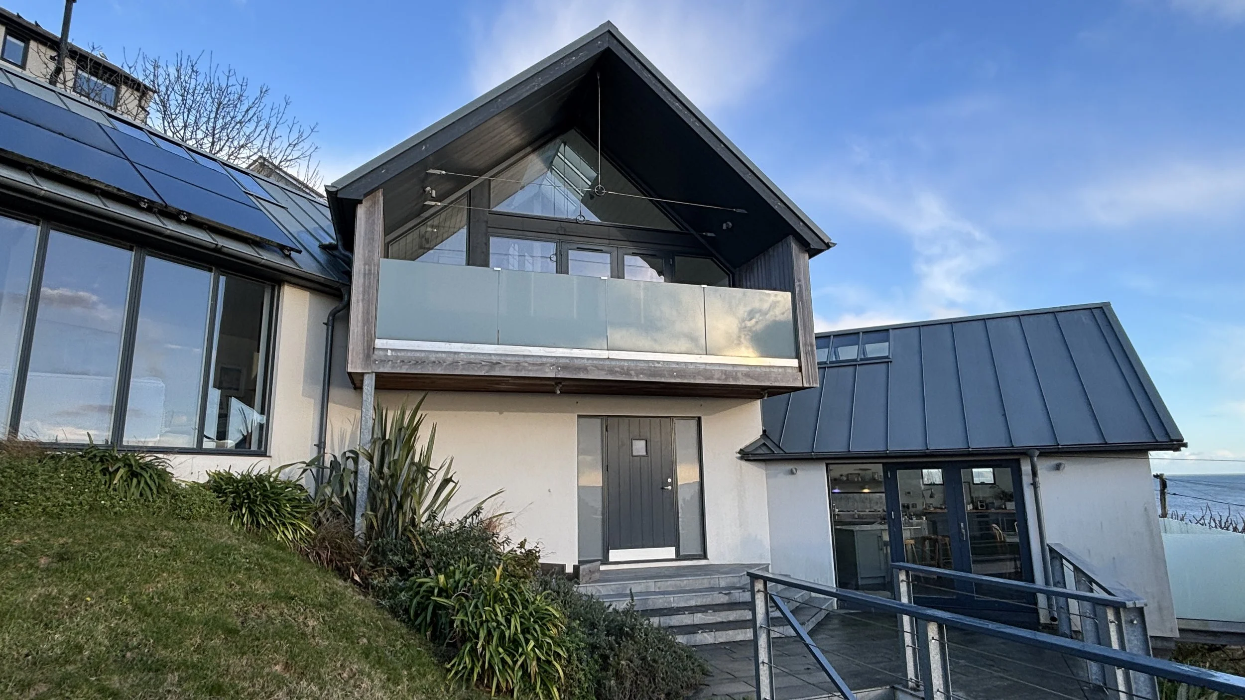 A modern house with a steep gable roof, large glass windows, and a balcony, situated on a sloped landscape with stairs and greenery.