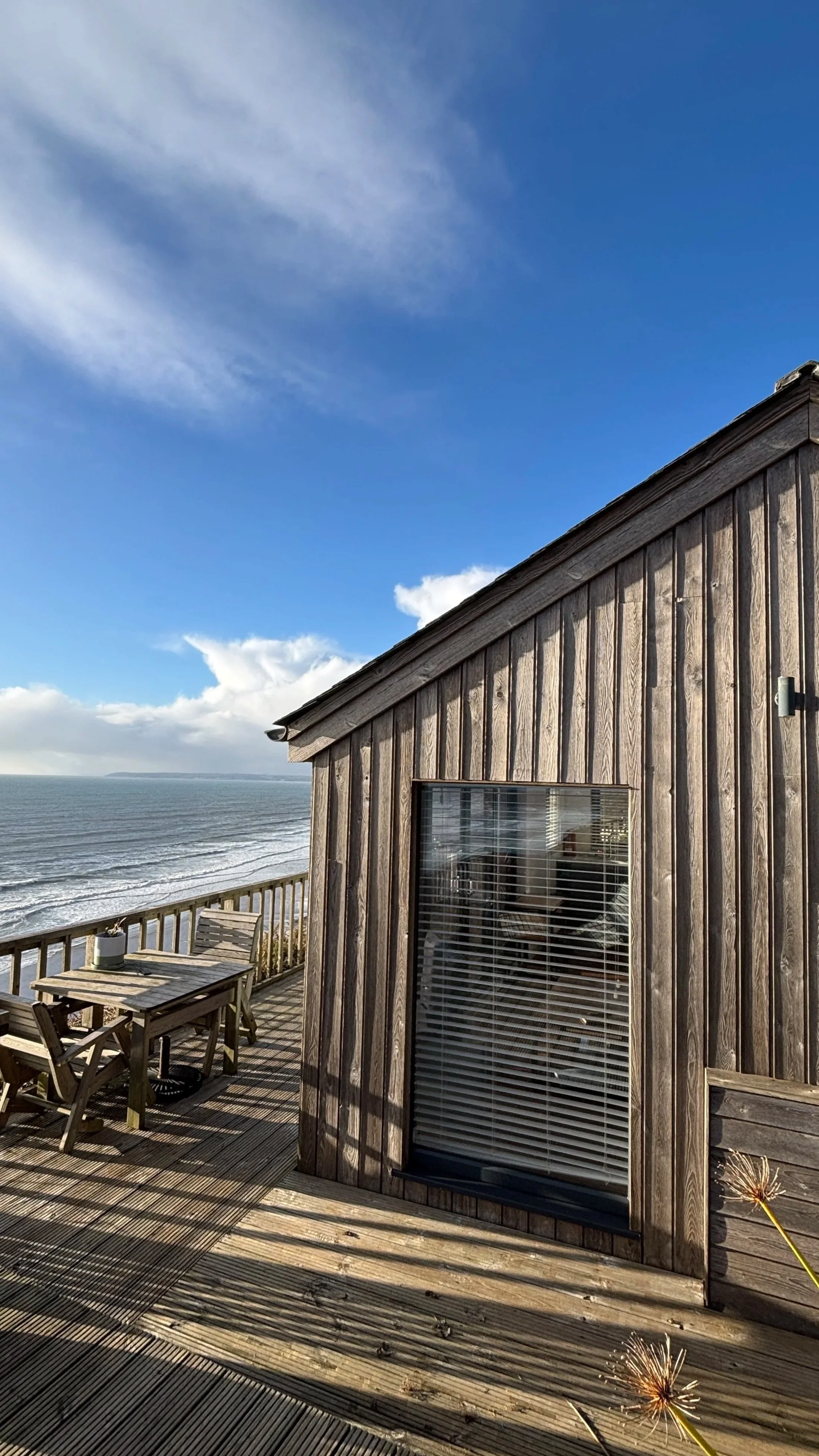 View of a wooden deck with outdoor furniture, overlooking the ocean with a partly cloudy sky.