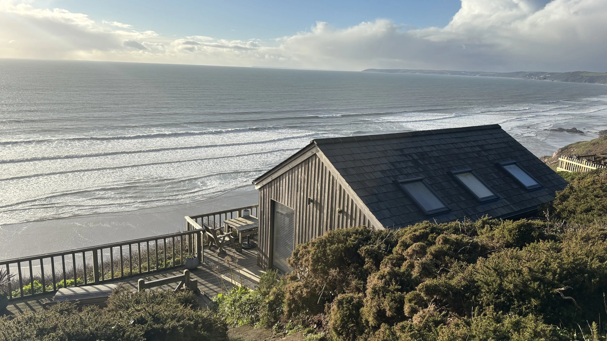 A view of the ocean with waves rolling onto the beach, seen from a wooden deck attached to a house with skylights, surrounded by bushes and greenery.