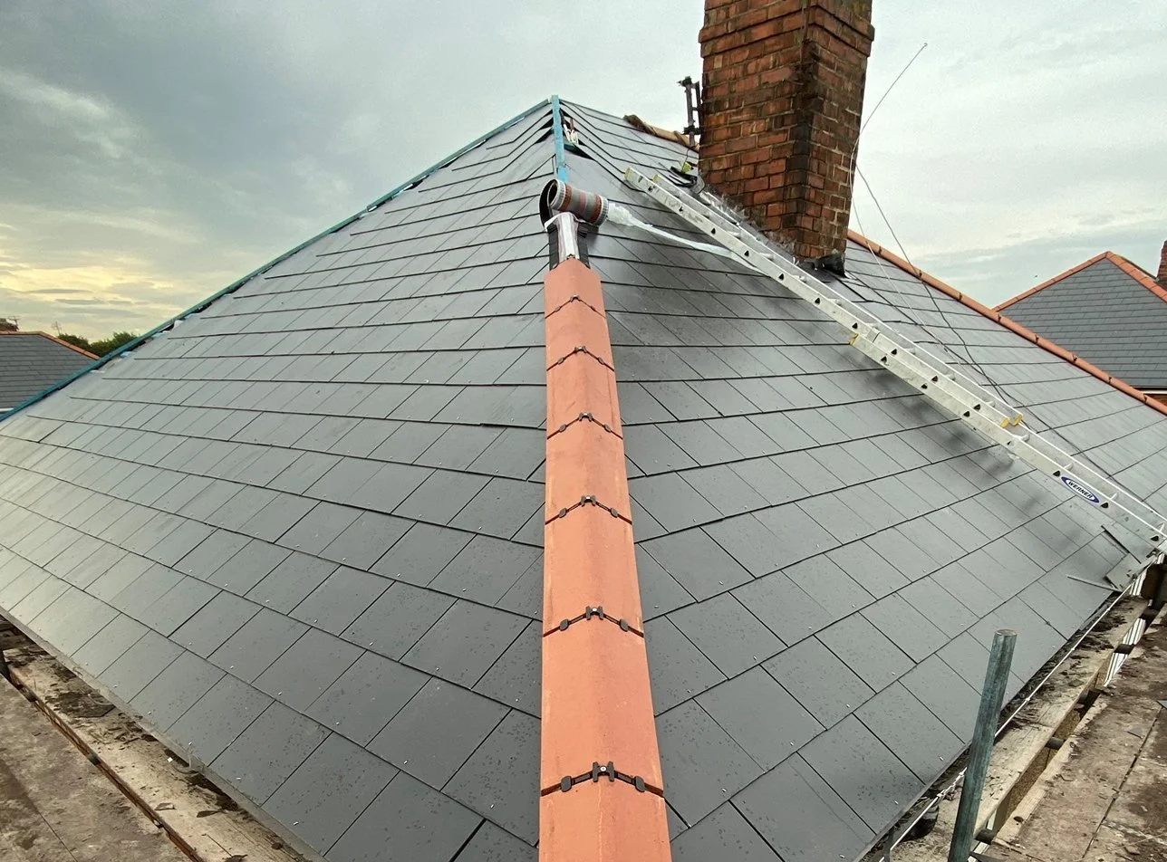 A rooftop under construction featuring gray tiles, a brick chimney, a ladder, and a vent pipe against an overcast sky.