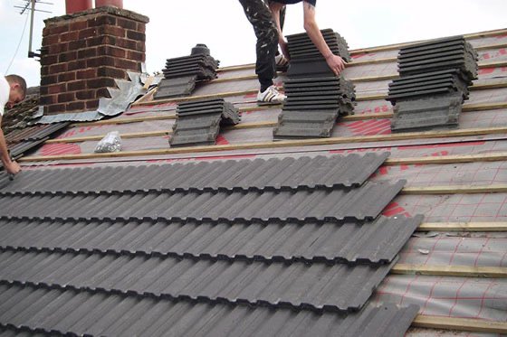 Person installing grey roofing tiles on a sloped roof with stacks of tiles and uninstalled sections visible.
