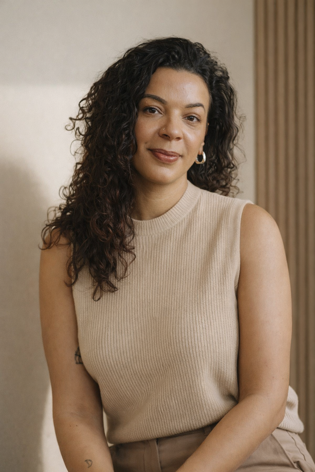 A woman with curly dark hair, wearing a sleeveless beige top, sitting in front of a neutral background with curtains.