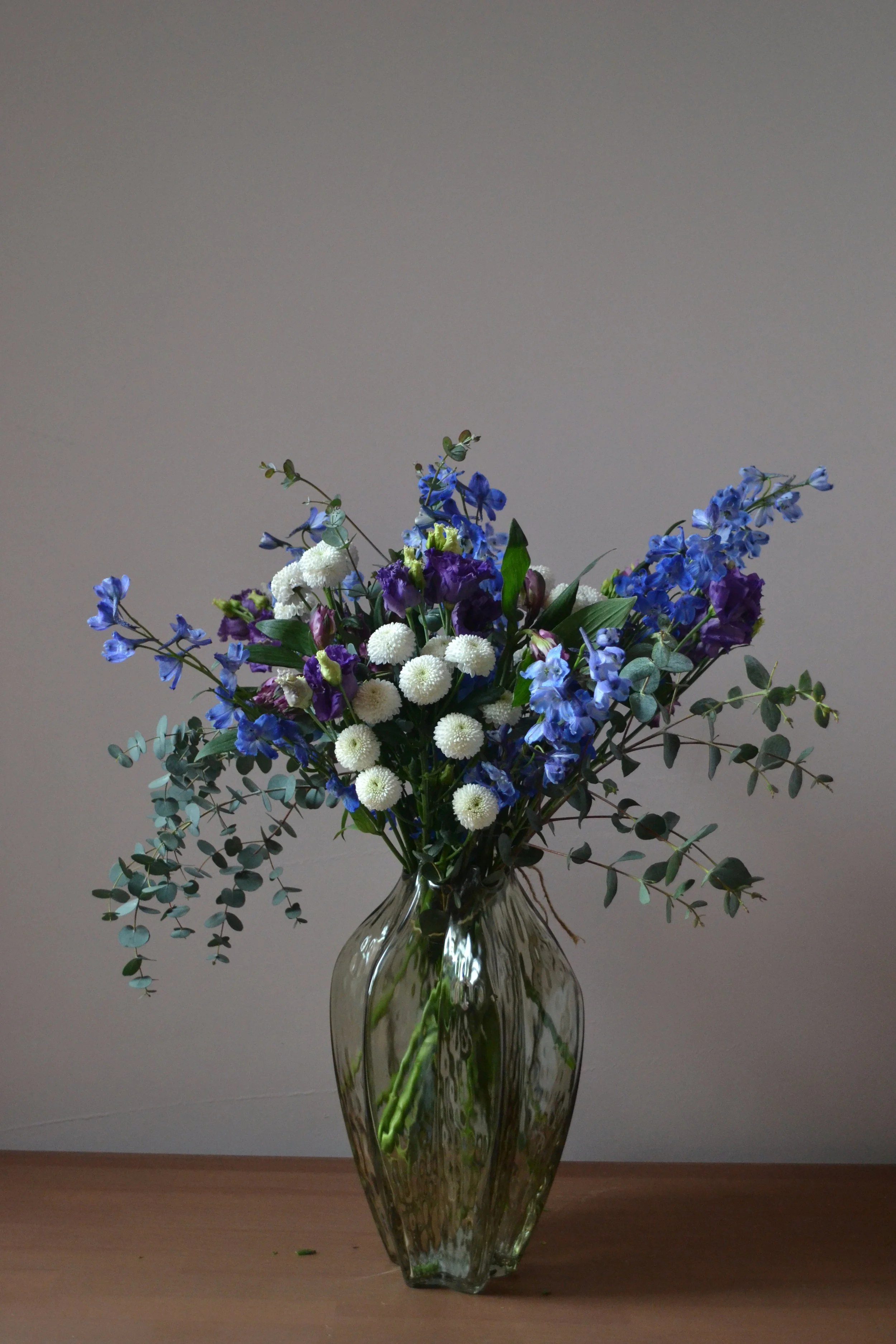 A glass vase with a bouquet of blue, purple, and white flowers, including greenery, placed on a wooden surface against a plain wall.