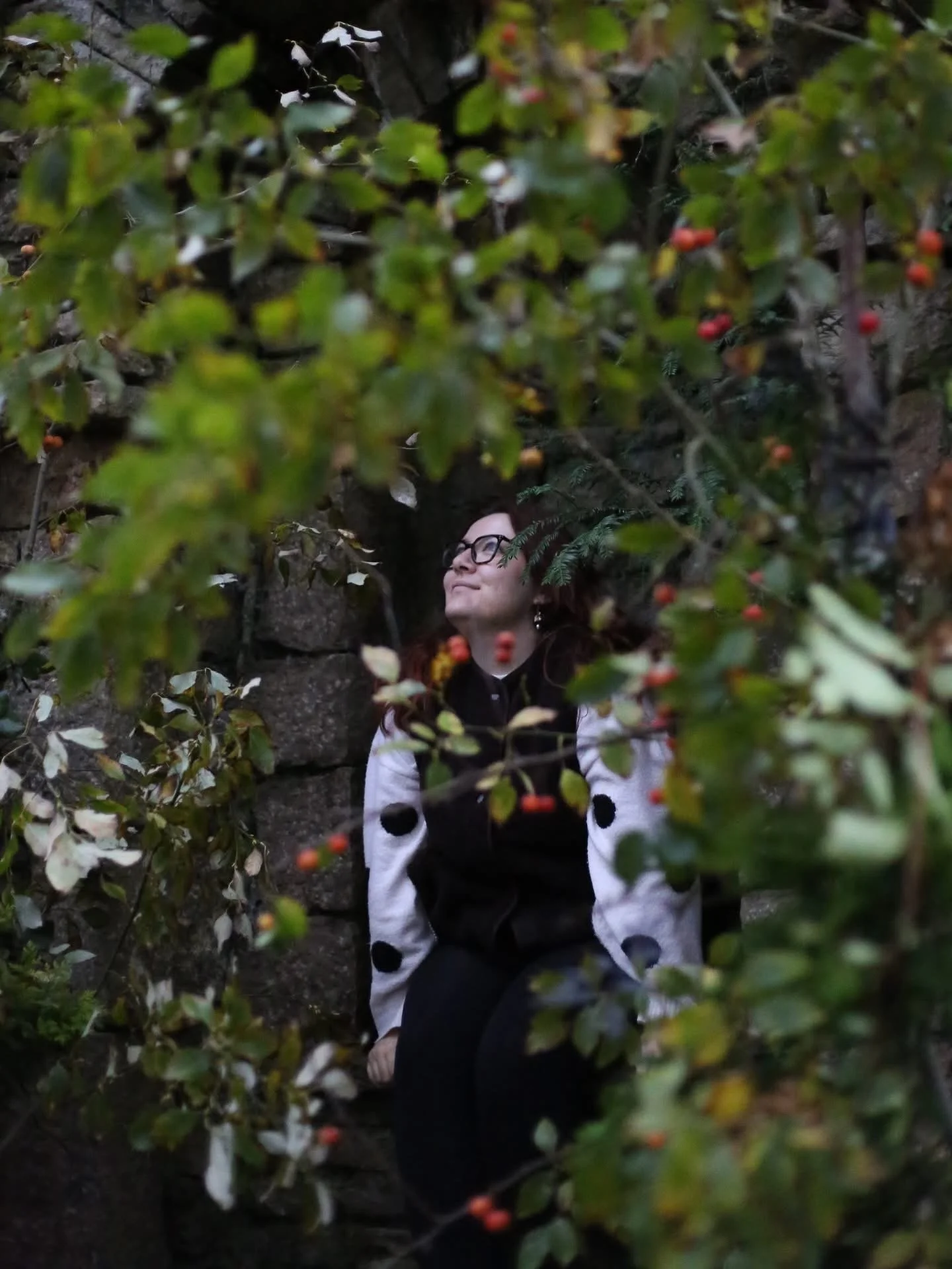 Woman with glasses and dark hair sitting among green foliage and berries, smiling with eyes closed.
