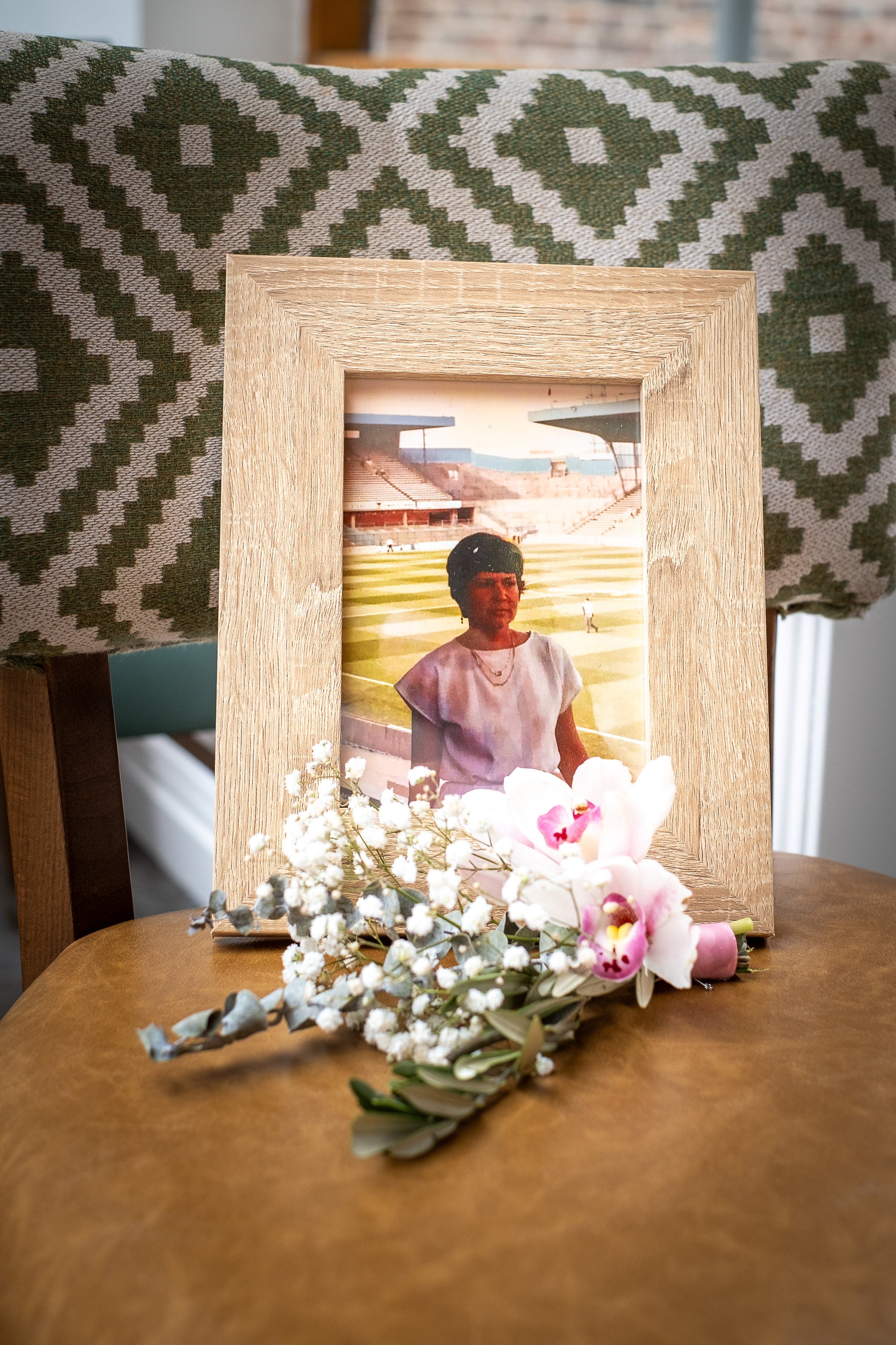 A framed photograph of a woman standing on a sports field, surrounded by flowers including white baby's breath and pink orchids, resting on a wooden surface with a patterned fabric backdrop.