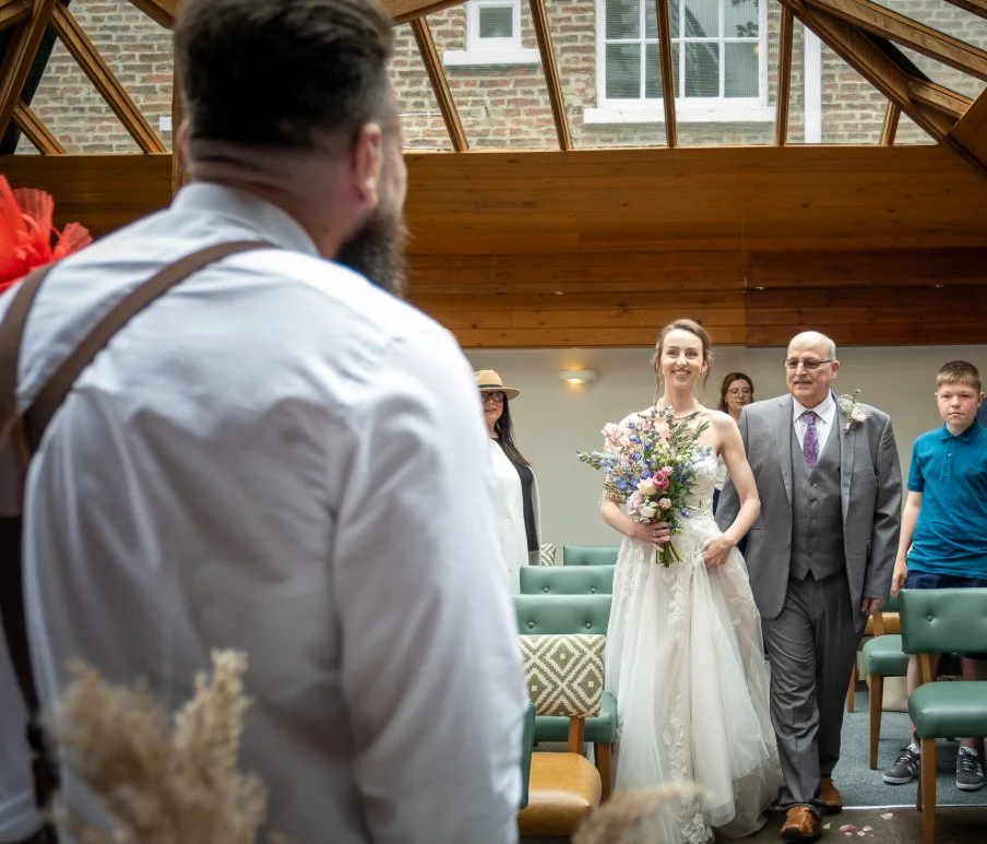 A bride walking down the aisle holding a bouquet of flowers, accompanied by a man, likely her father, inside a bright indoor space with a glass ceiling and wooden walls. Guests are seated on green chairs, watching the bride.