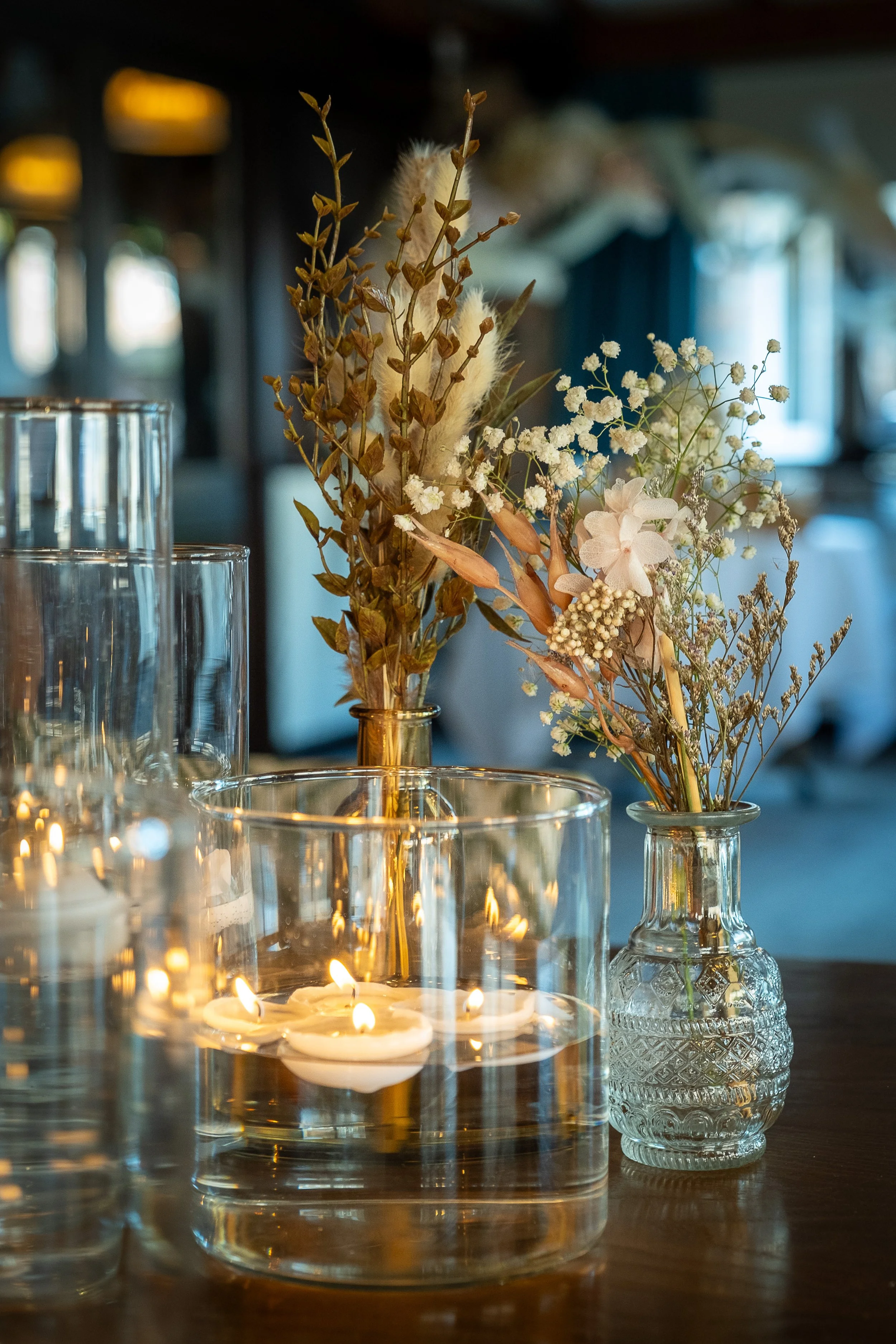Decorative arrangement of dried flowers in glass vases and lit candles on a wooden table.