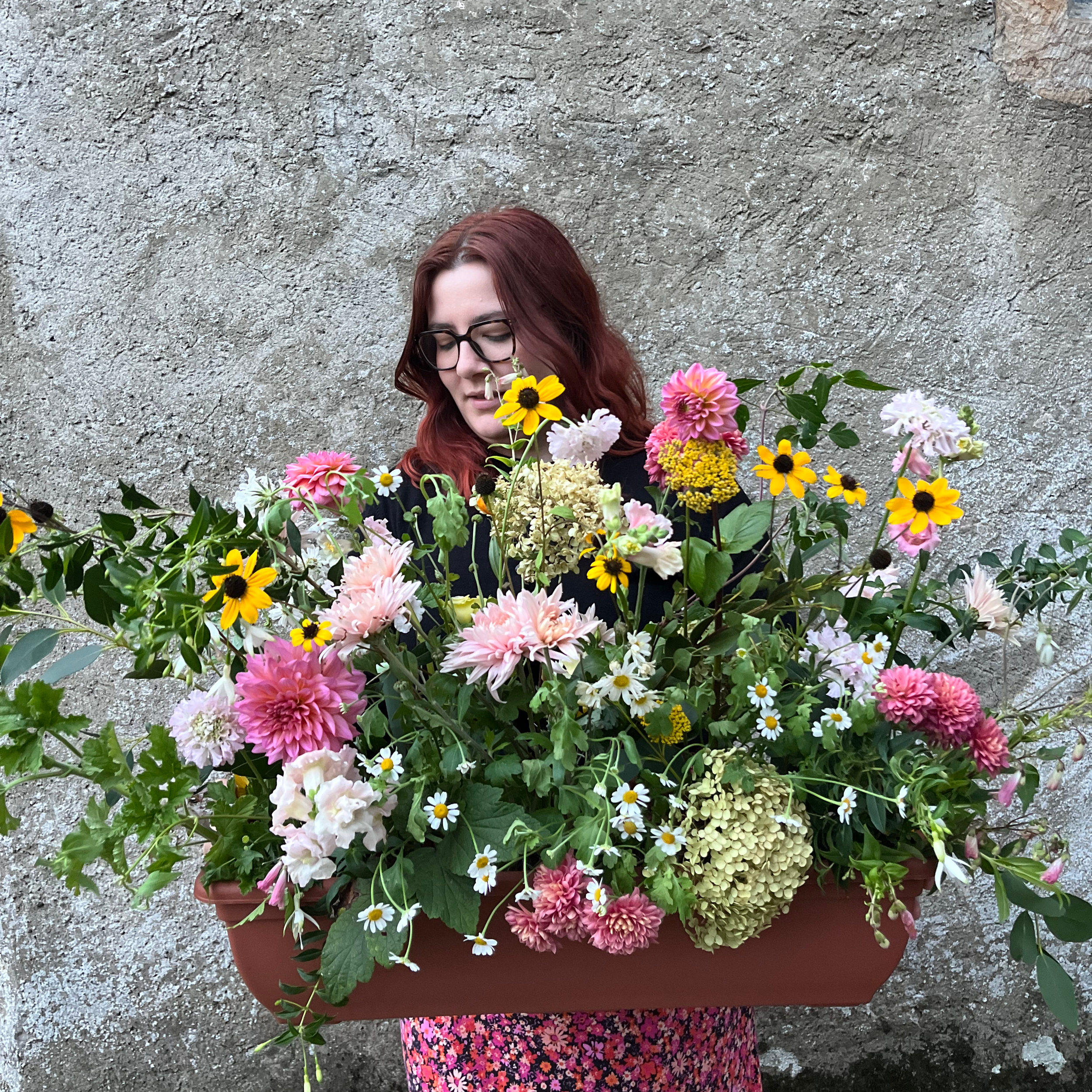 A woman with red hair and glasses holding a large tray of colorful flowers in front of a textured wall.