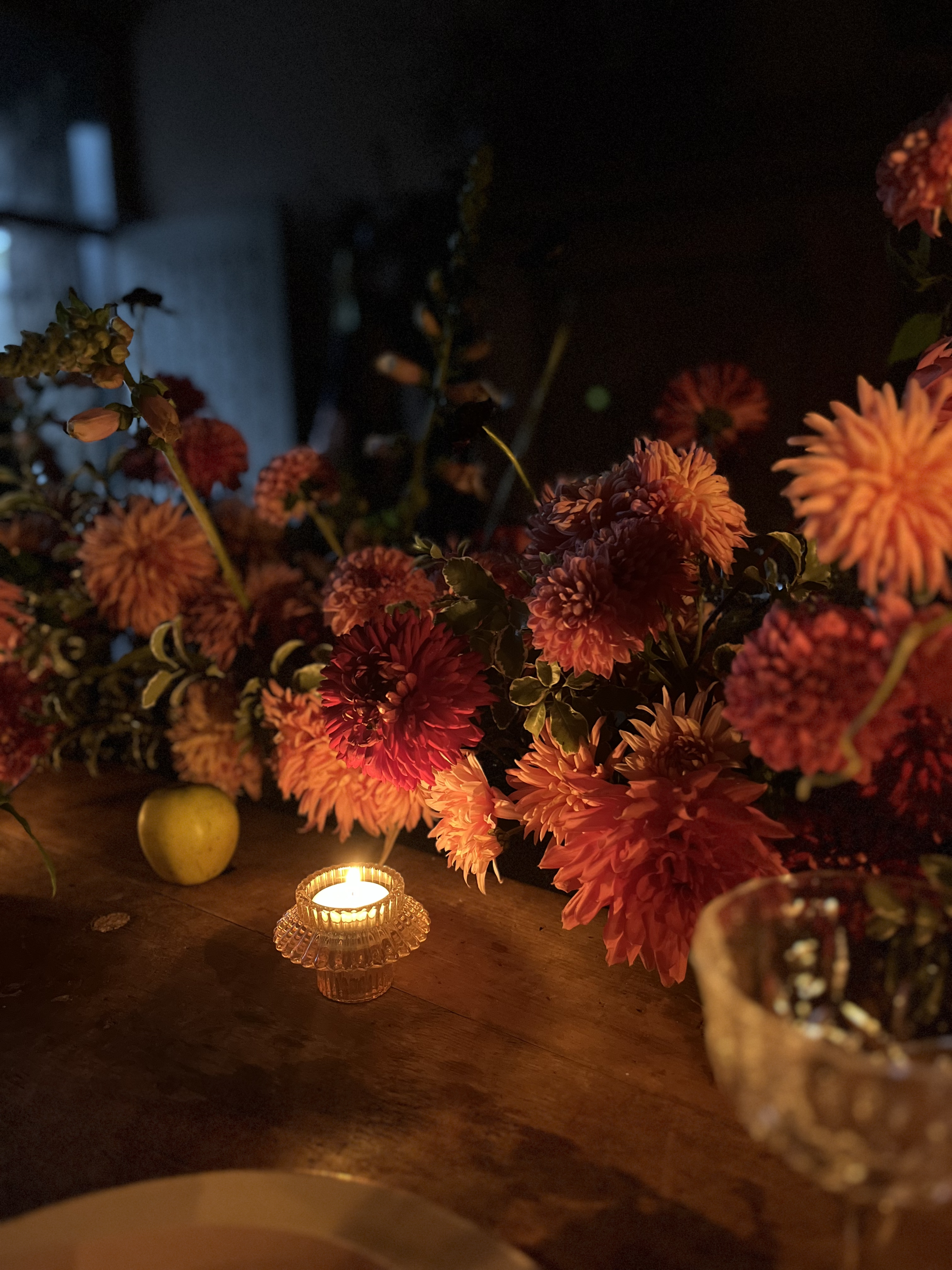 Vase of pink and orange flowers, yellow apple, lit candle in a glass holder on a wooden table.