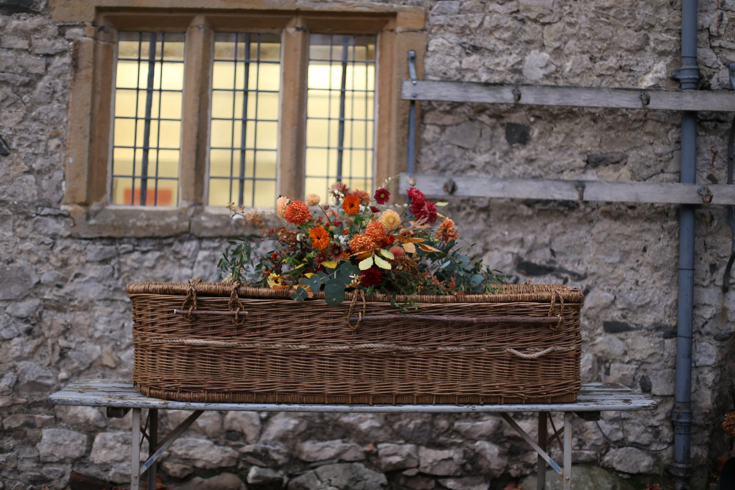 A rectangular woven basket filled with orange and red flowers placed on a weathered wooden table, with a stone wall and barred window in the background.