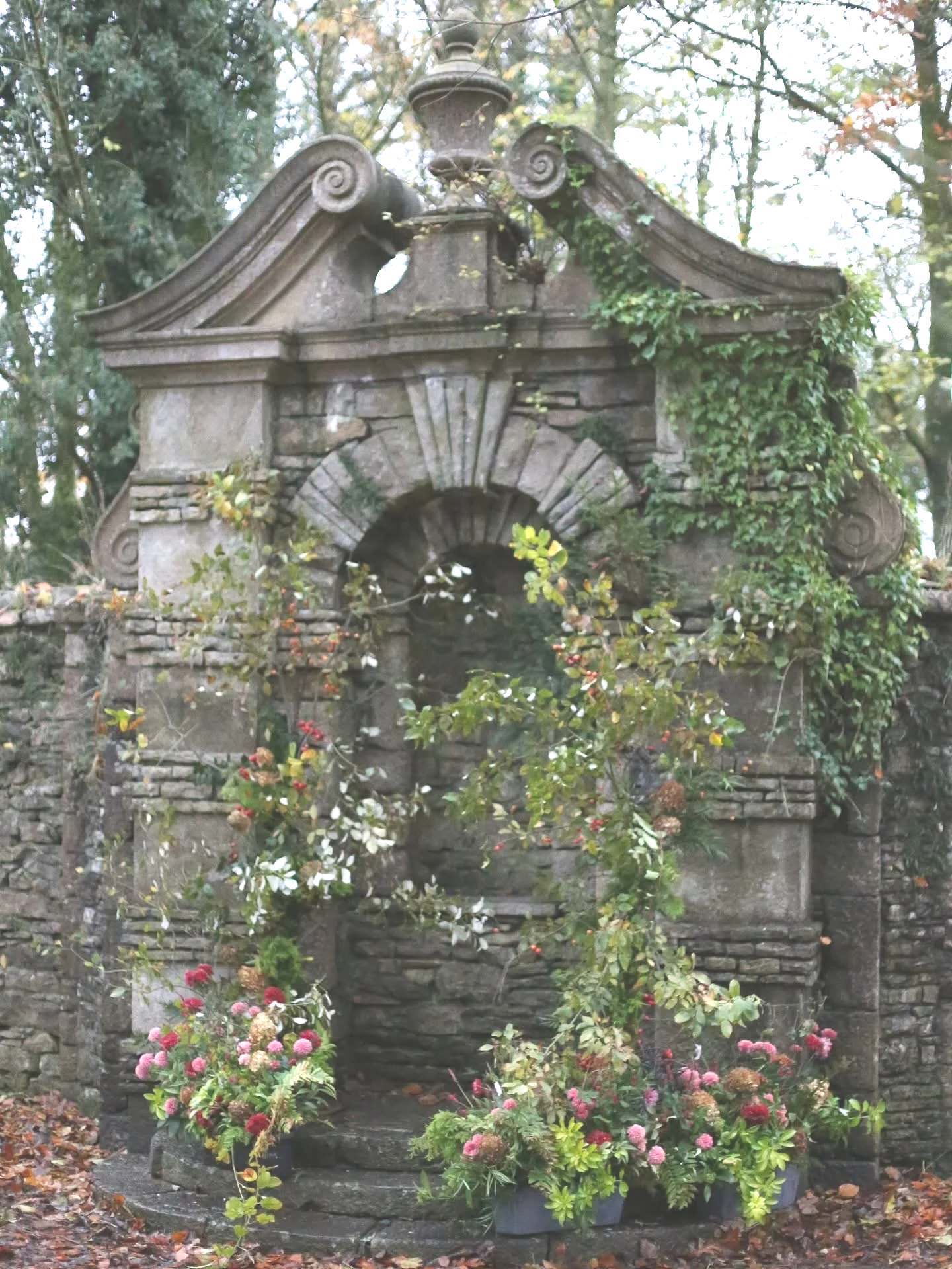A stone fountain with a decorative top, surrounded by greenery and flowering plants in pots.