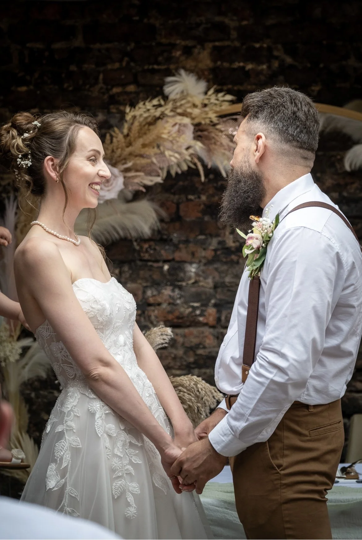 A bride and groom holding hands during their wedding ceremony, smiling at each other, with a rustic brick wall and decorative dried flowers in the background.