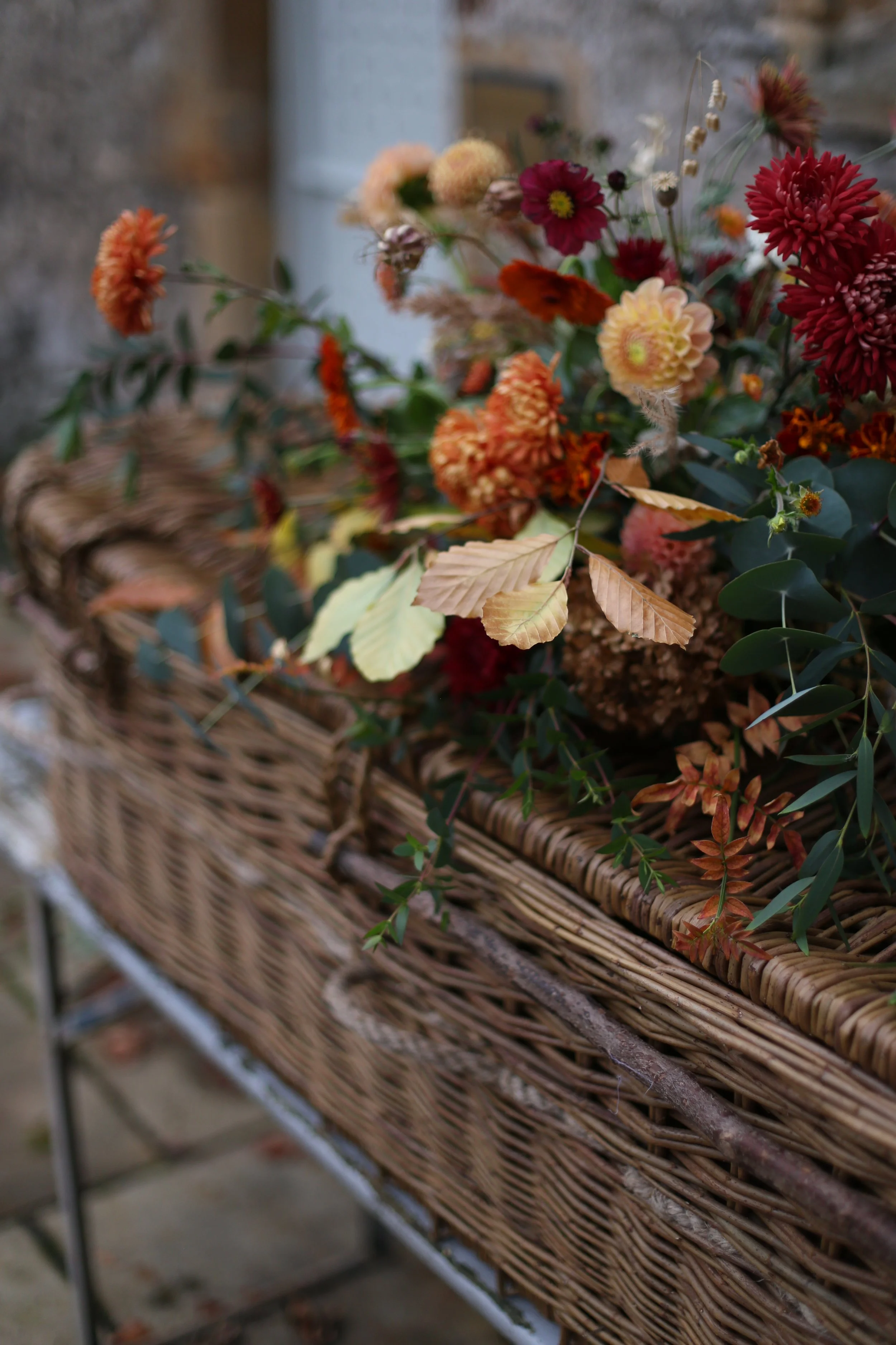 A wicker basket filled with fall-colored flowers and leaves, set outdoors on a stone surface.