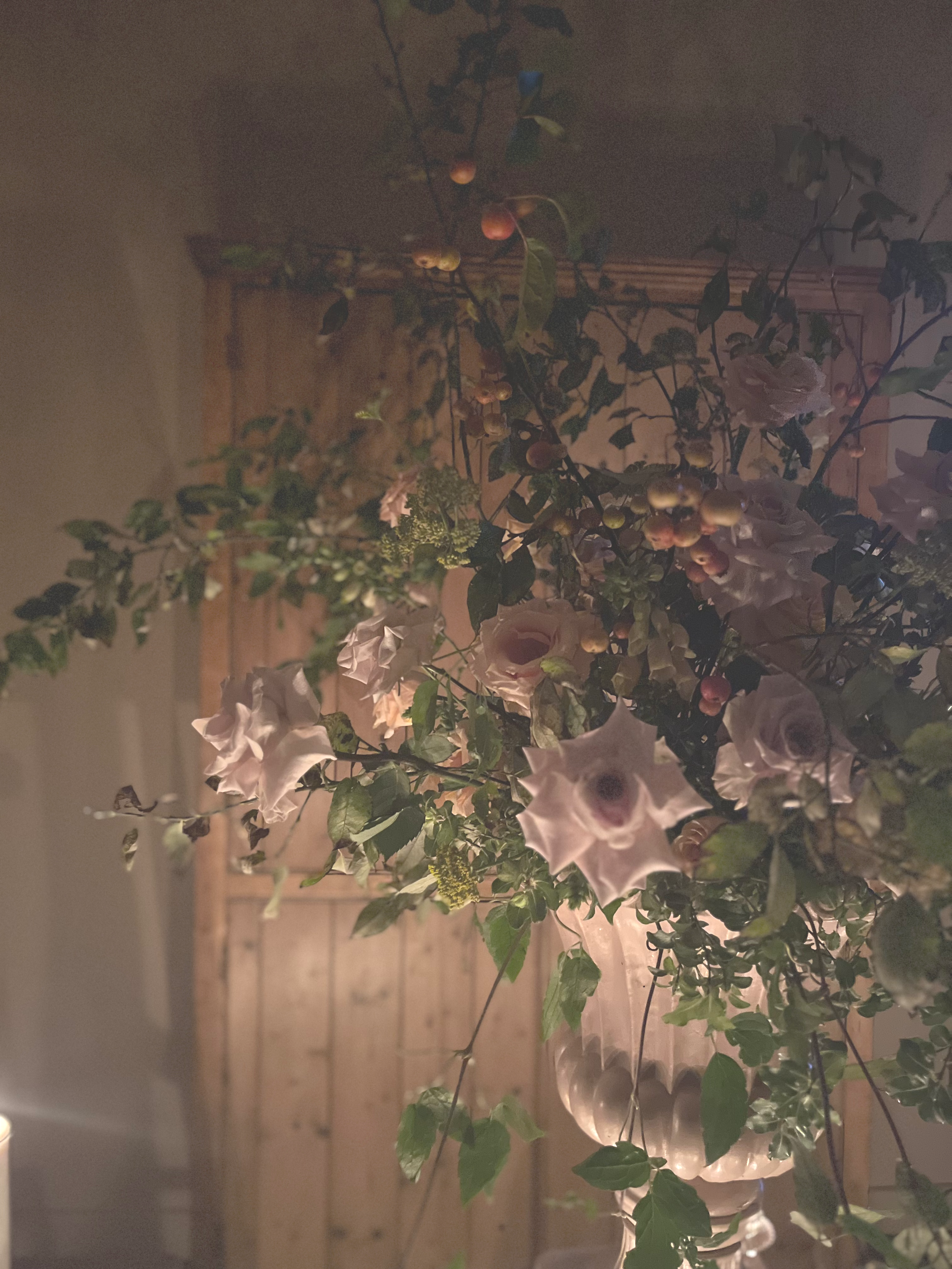 A floral arrangement with white and pink flowers, green leaves, and small pink and green buds in a decorative vase, set against a wooden wall background.