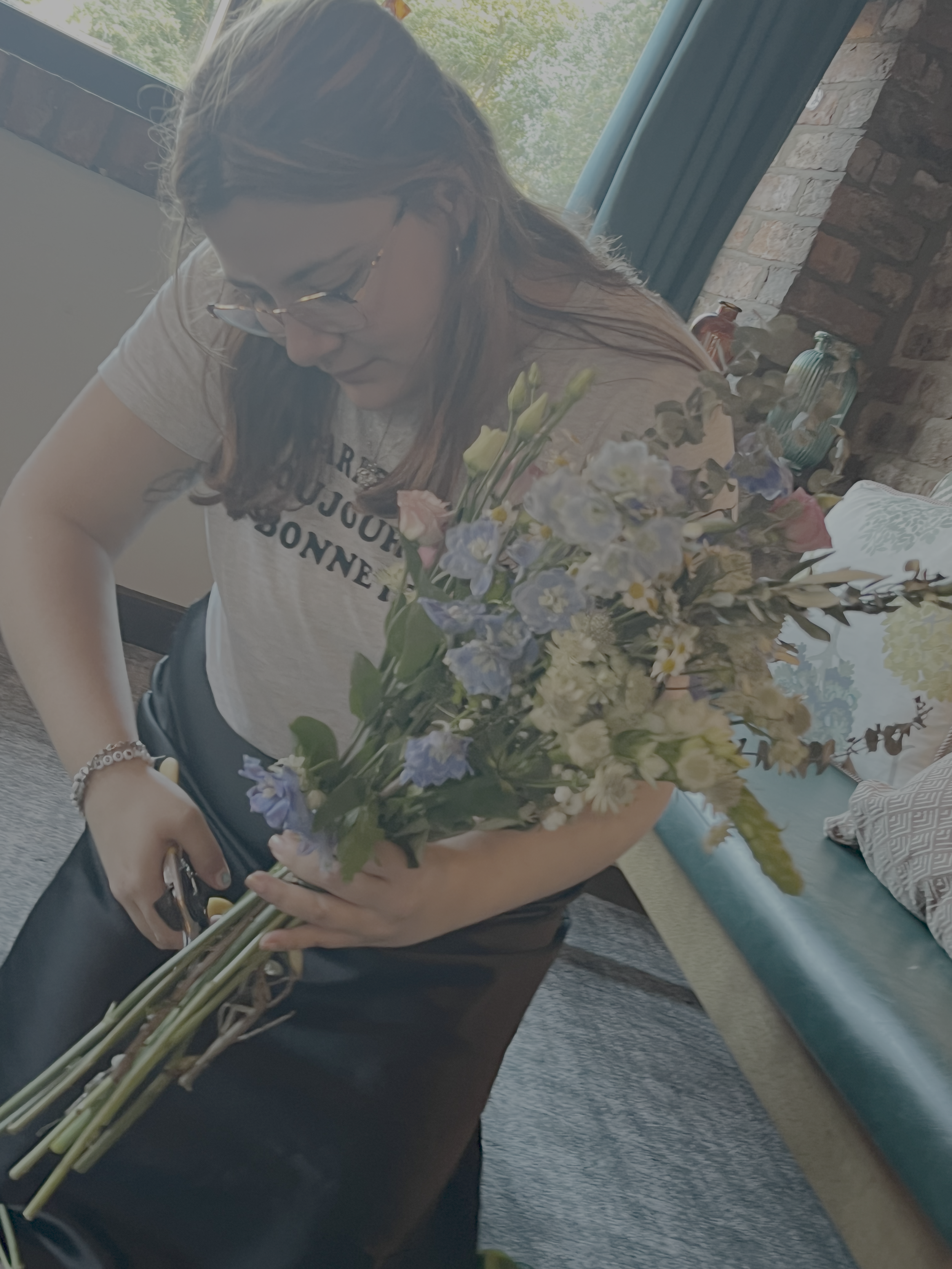 A woman with glasses arranging a bouquet of fresh flowers with various colors including purple, white, and yellow, in a cozy room with a blue sofa and a brick wall in the background.