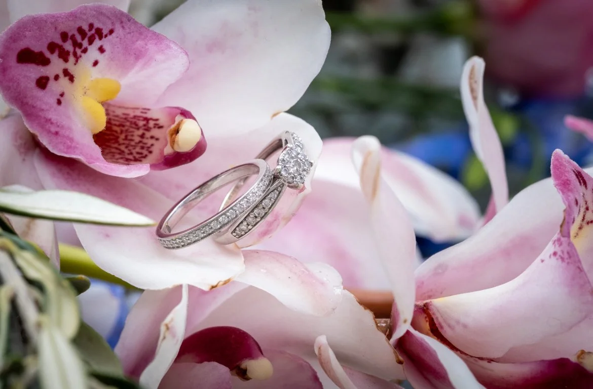 Two rings, one with a large central diamond and smaller surrounding diamonds, resting on pink and white orchid flowers.