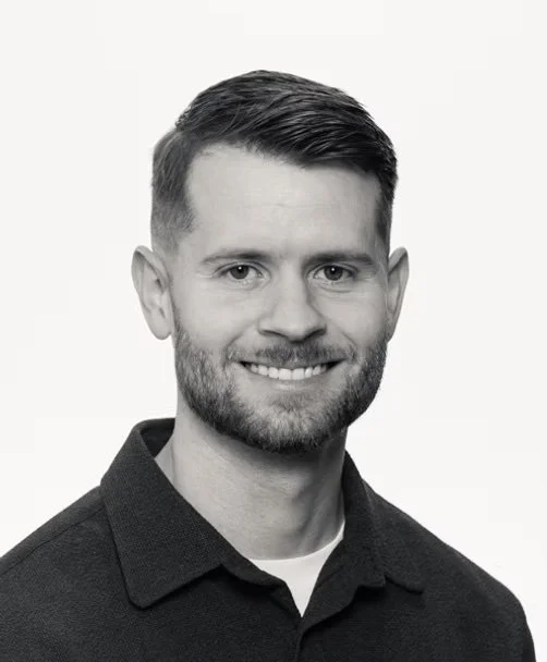 Black and white headshot of a smiling man with short, dark hair and a beard wearing a dark collared shirt.