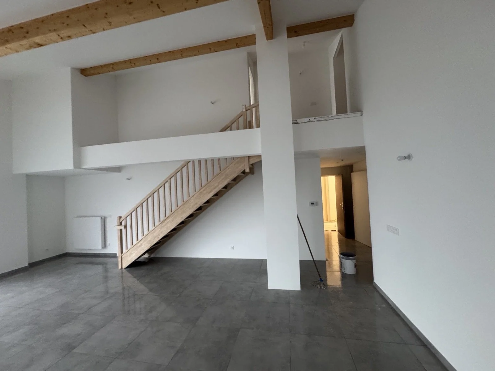 Intérieur d'une maison neuve avec murs blancs, sol en carrelage gris, escalier en bois menant à un étage, et un couloir lumineux.