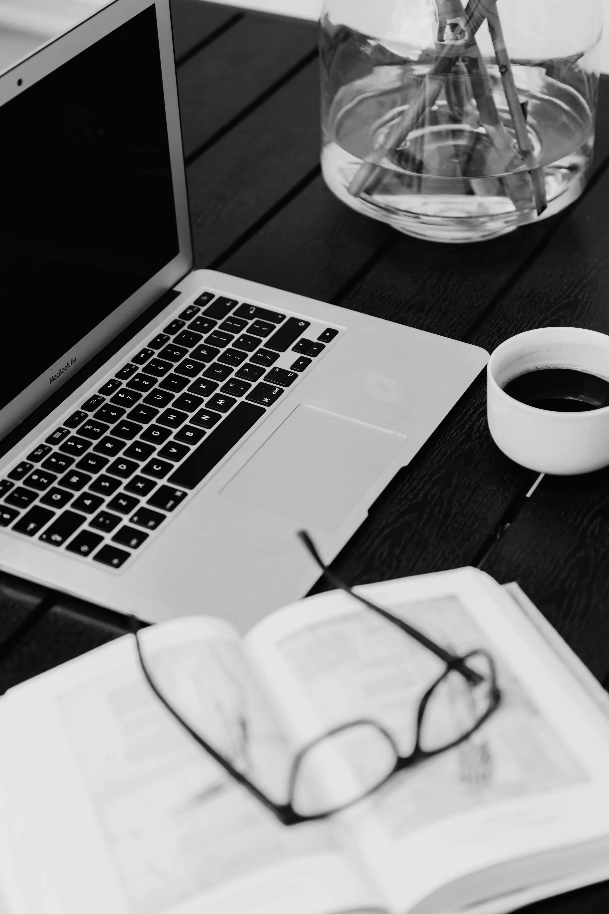 Un bureau avec un ordinateur portable, une tasse de café, un livre ouvert avec des lunettes et un vase avec des crayons.