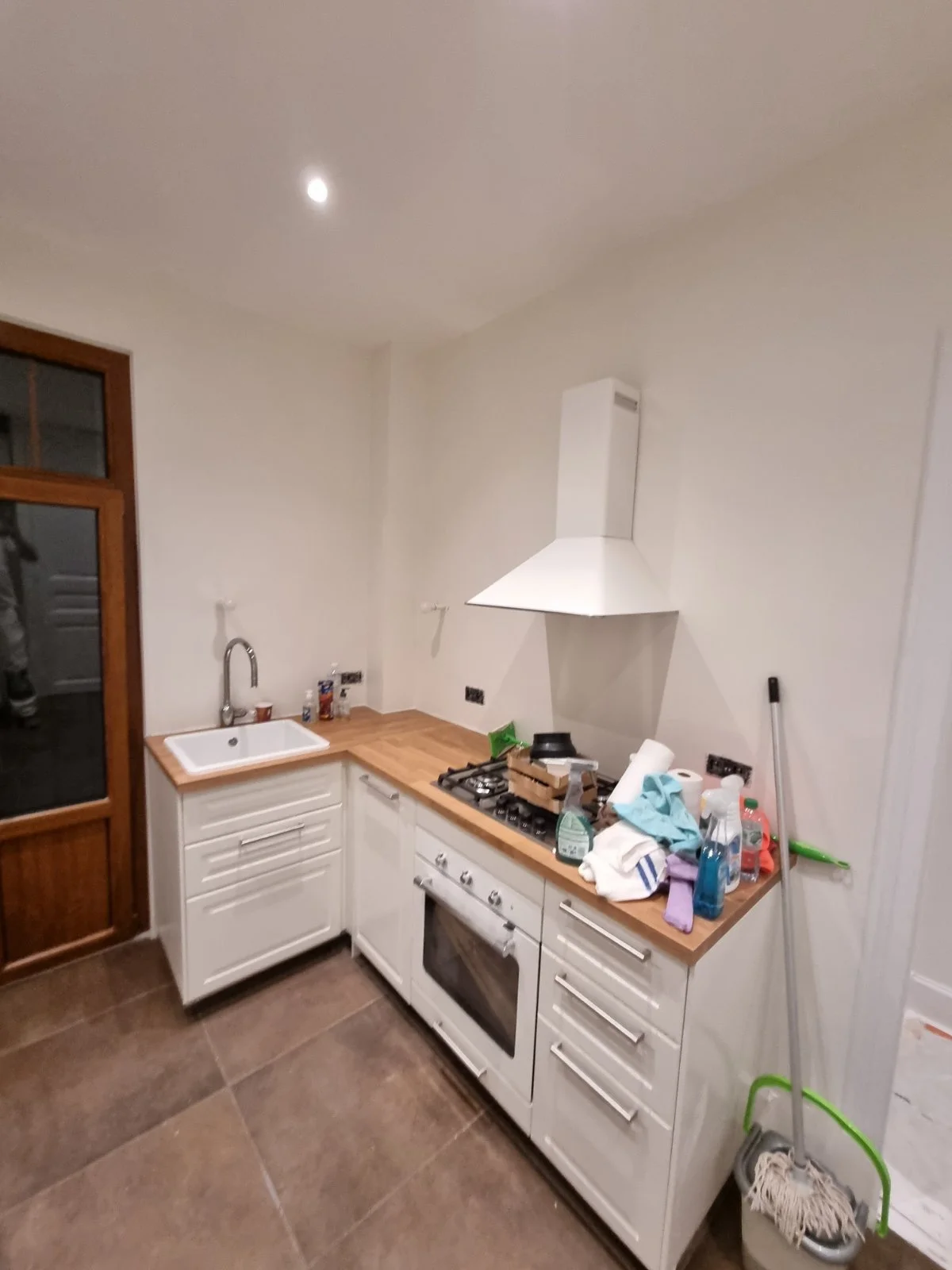 Cuisine avec un évier, une cuisinière à gaz, un plan de travail en bois, et divers objets de nettoyage et vaisselle sur le plan de travail.