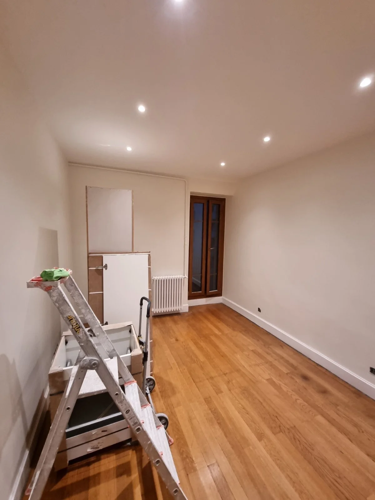 Une pièce vide en cours de rénovation avec un plafond éclairé, un radiateur, un escalier de servante, et un parquet en bois, avec une porte-fenêtre.