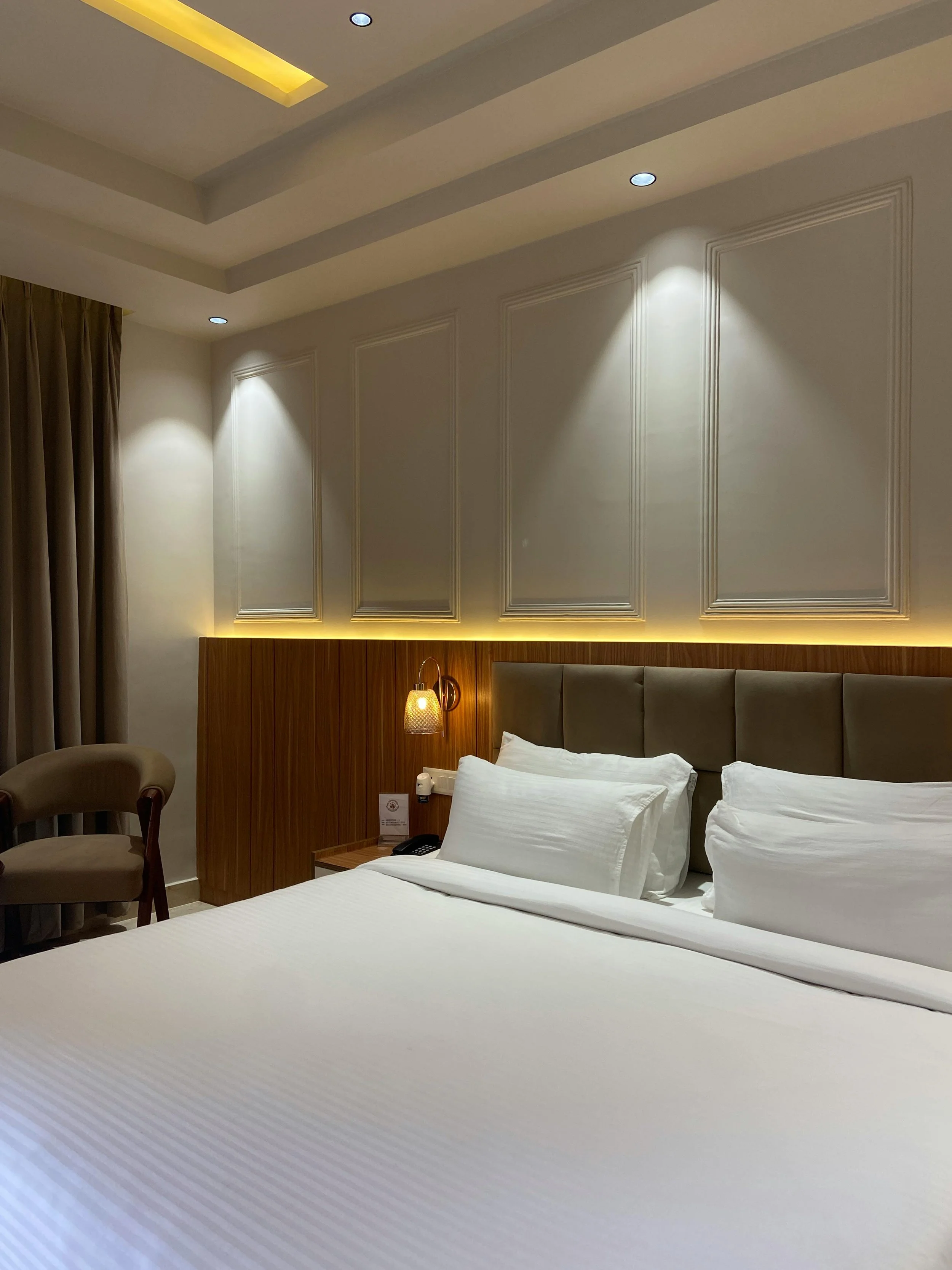 Chambre d'hôtel avec un lit double, tête de lit rembourrée, lampadaire adjacent, mur en panneaux décoratifs, et chaises dans un coin, avec lumière tamisée.