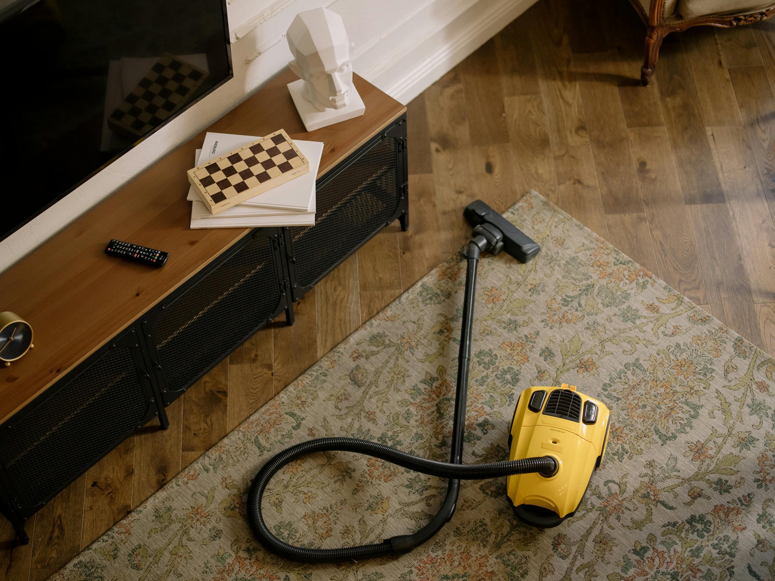 Un aspirateur jaune posé sur un tapis beige à motifs, dans une pièce avec un sol en bois, à côté d'une console en bois contenant une télé, des livres, un bâton de jeu d'échec et une sculpture blanche.