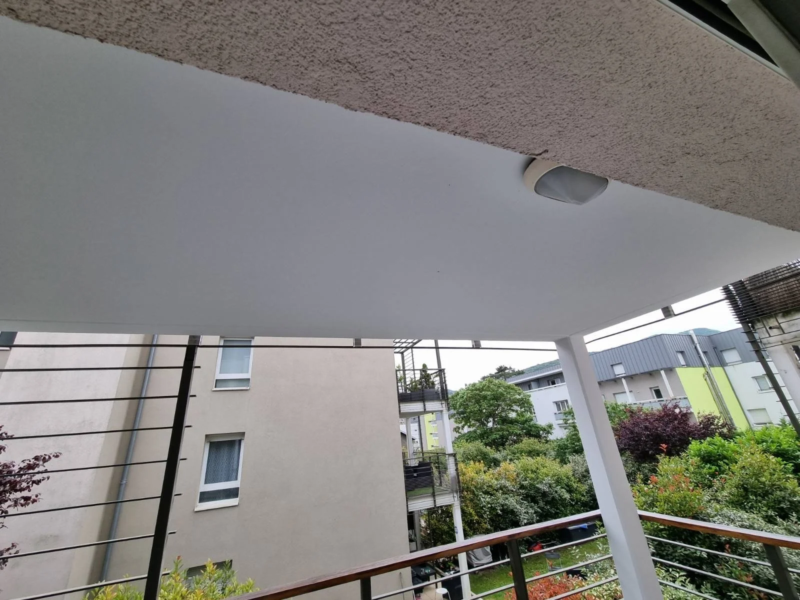 Vue depuis un balcon montrant une toiture, un plafond blanc avec un luminaire, des bâtiments modernes et un espace vert avec des arbres et des buissons.