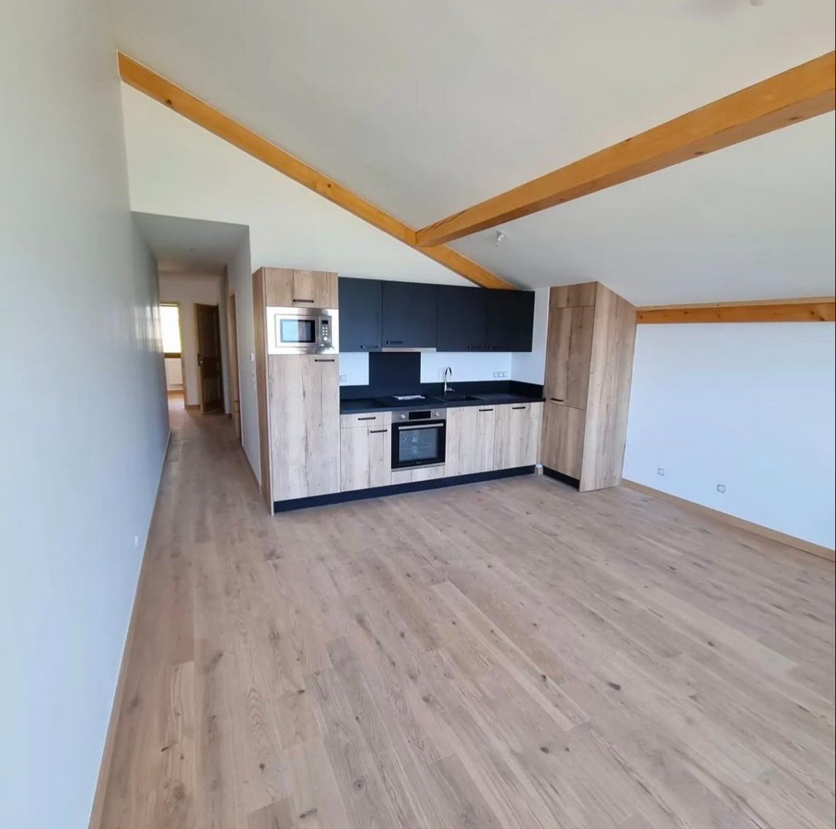 Cuisine moderne dans un appartement avec mur en bois, plafond en pente avec poutres en bois, et sol en parquet. La cuisine est équipée d'armoires en bois clair, d'un four, d'une plaque de cuisson, d'un évier et d'un micro-ondes encastré.