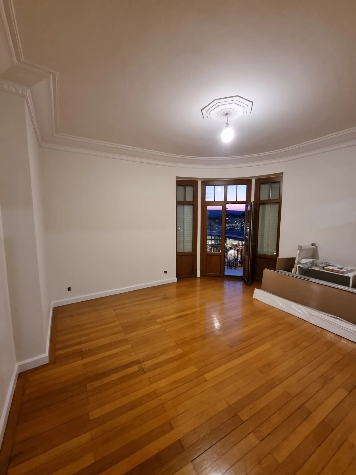 Pièce vide avec parquet en bois clair, grande fenêtre avec porte-fenêtre en bois ouvrant sur un balcon, plafond avec moulures et lampe suspendue.