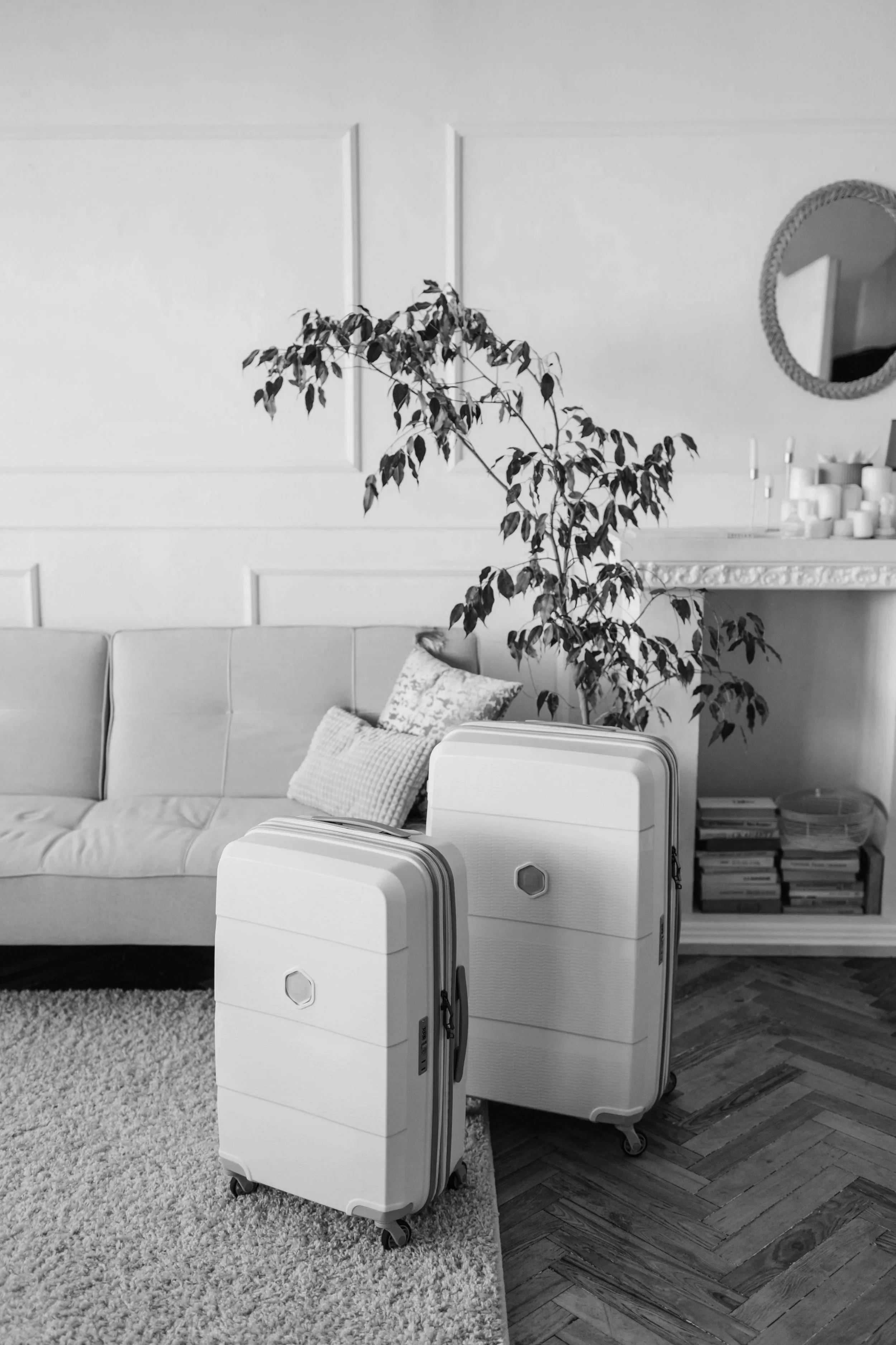Deux valises blanches à roulettes dans un salon moderne avec un canapé gris, des coussins, une plante d'intérieur, un miroir rond, une cheminée décorative et une étagère remplie de livres.