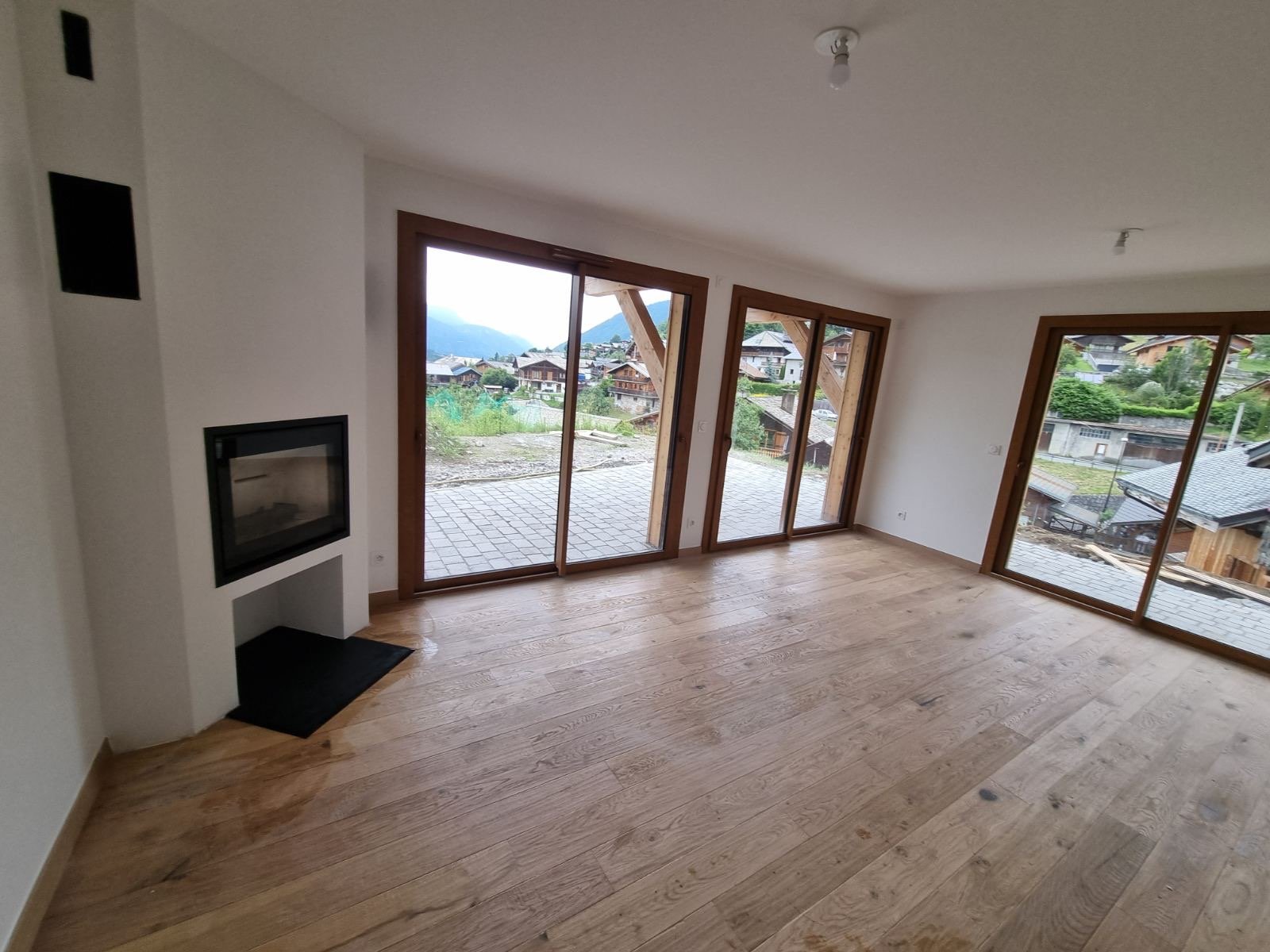 Salon vide avec parquet en bois clair, grandes fenêtres en bois offrant une vue sur un village de montagne, cheminée encastrée dans un mur blanc, plafonds avec lampes spot, vue extérieure avec maisons en bois et montagnes.