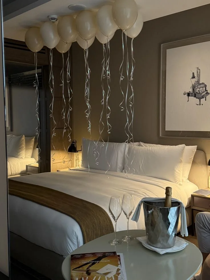 Chambre d'hôtel décorée pour une célébration avec des ballons blancs suspendus au plafond, bouteille de champagne dans un seau à glace avec deux flûtes, et une table avec un magazine et un élément de décoration.