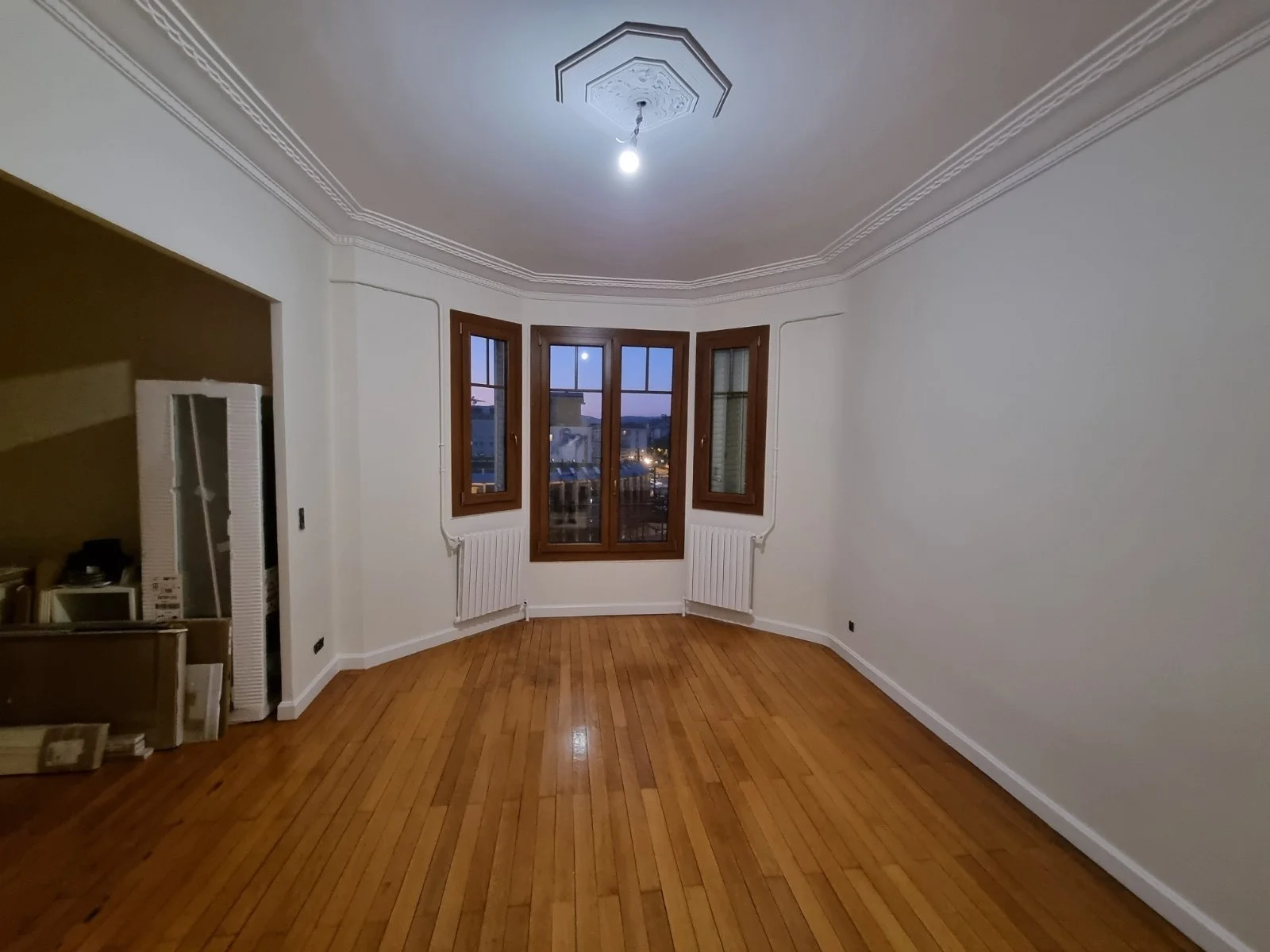 Une pièce vide avec des murs blancs, un parquet en bois clair, trois fenêtres en bois avec vue sur la ville, un plafond orné avec un luminaire suspendu, et un espace avec des cartons et une étagère.