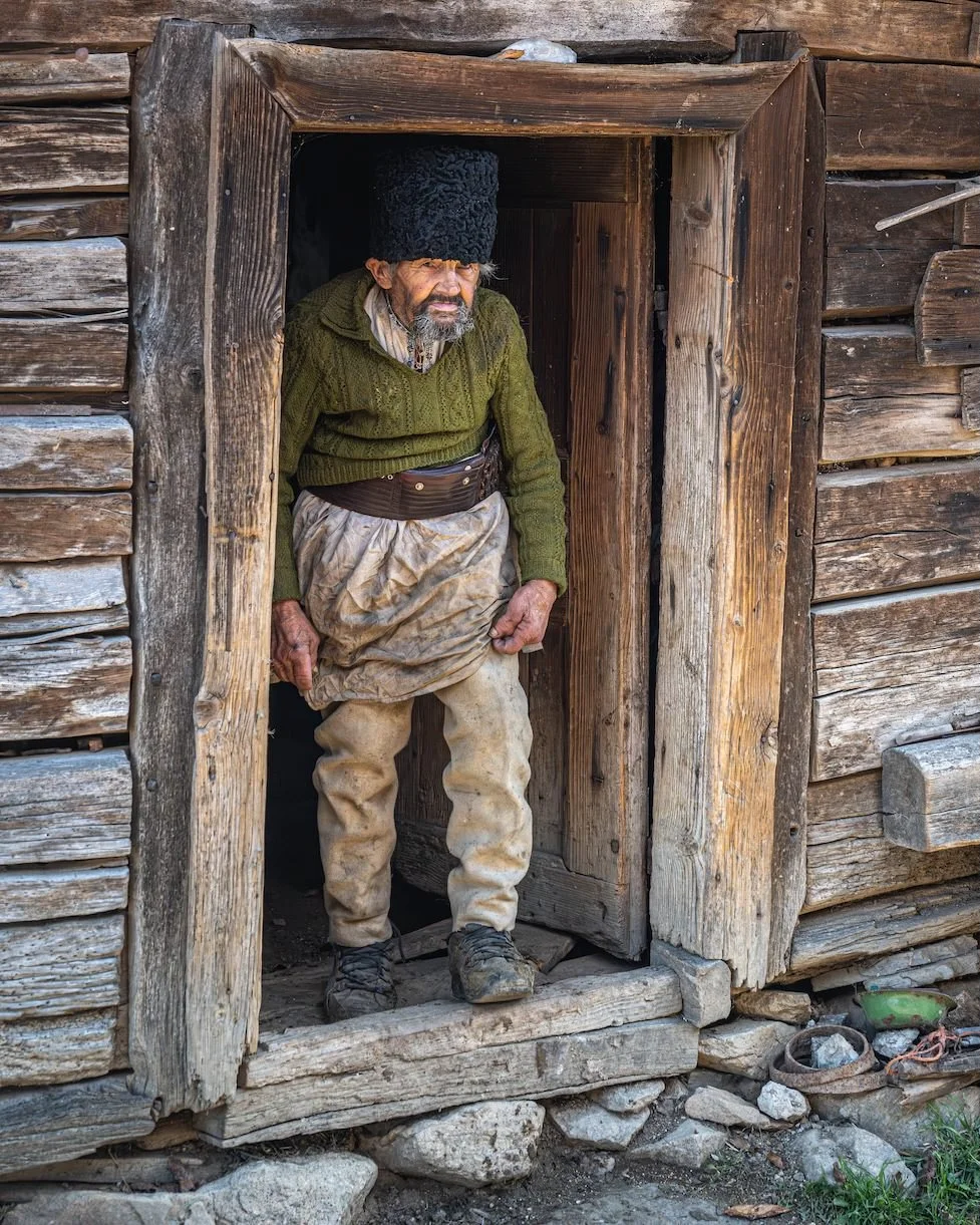An elderly man standing at a wooden doorway of a rustic cabin, wearing traditional clothing including a tall black hat, green sweater, and dirty beige pants.