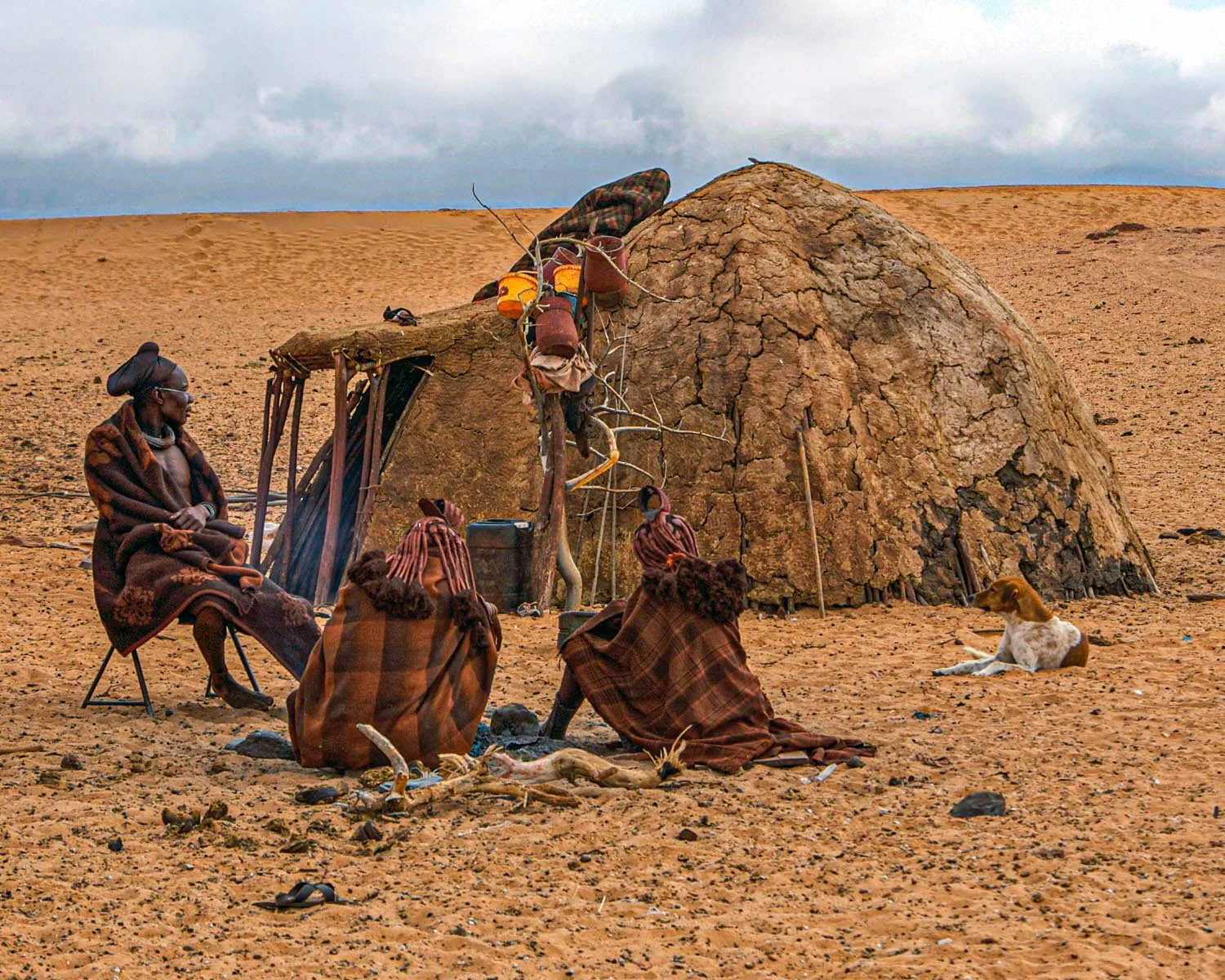 A group of indigenous people sitting outside a traditional mud hut in a desert landscape, with a dog lying on the ground nearby, under a cloudy sky.