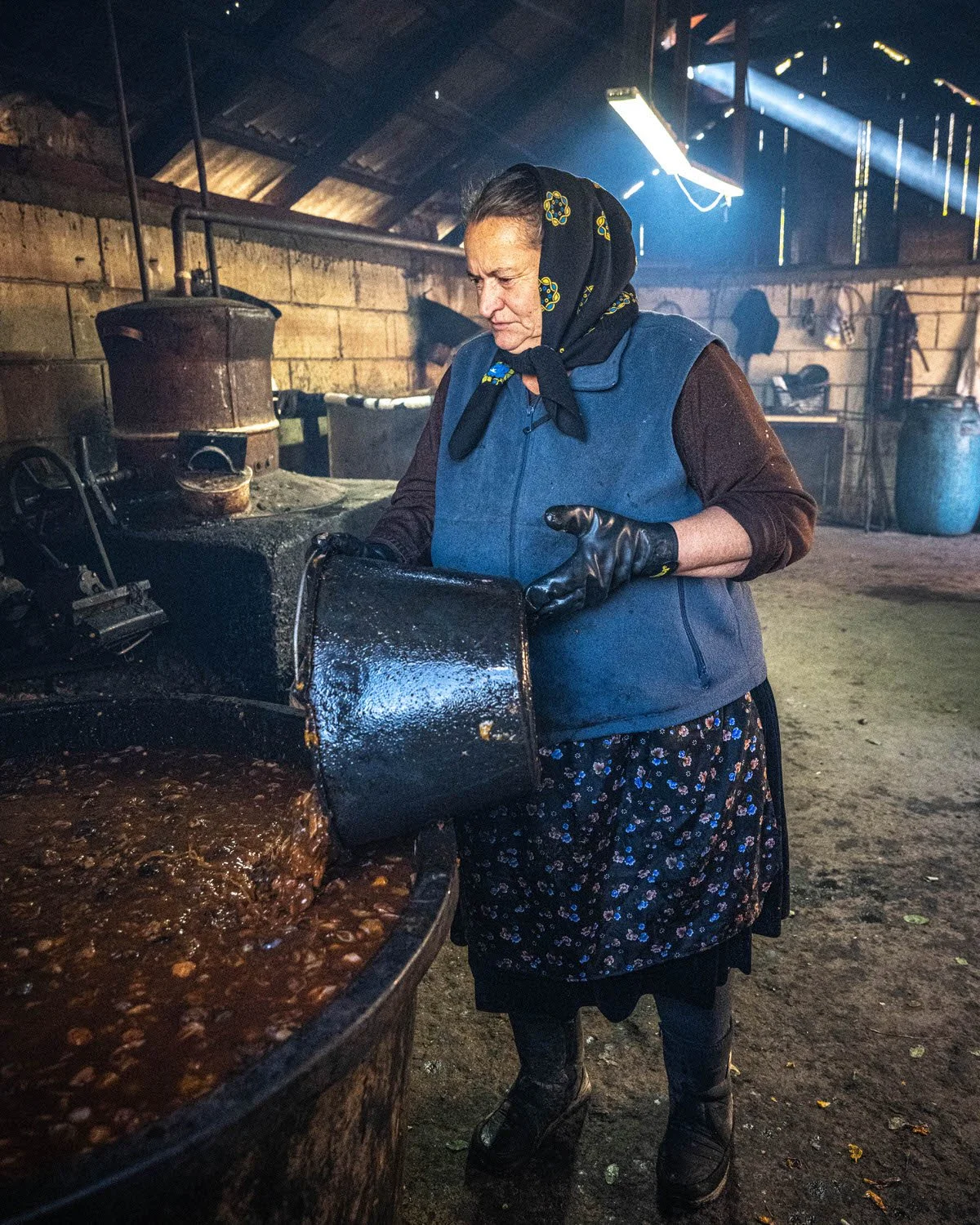 An elderly woman wearing a headscarf, dark gloves, a blue vest, and a floral skirt, pouring a substance into a large cauldron of boiling food inside a rustic, dimly lit barn or kitchen.