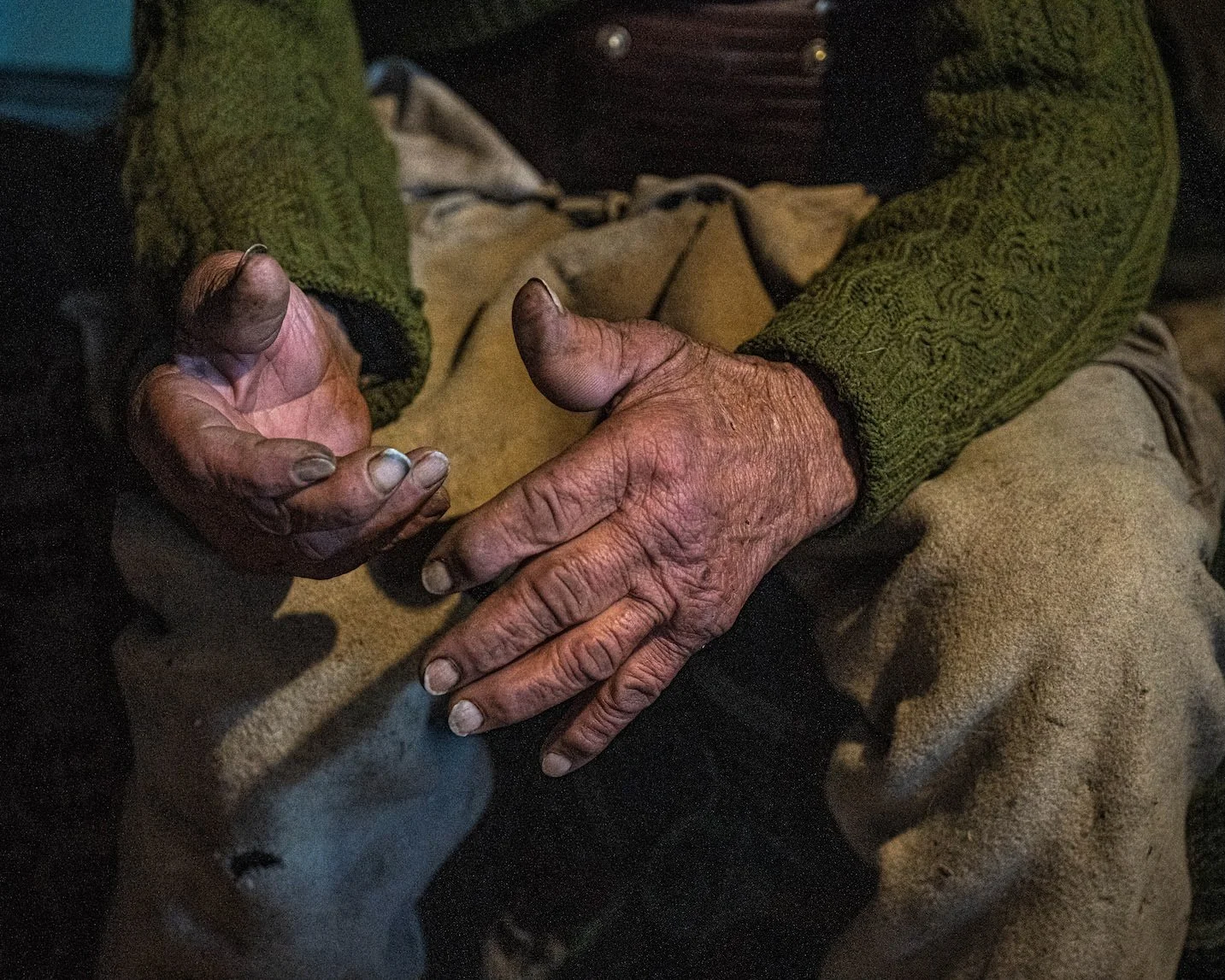 Close-up of elderly person's hands, one reaching out to touch another hand, with worn clothing and a textured green jacket.