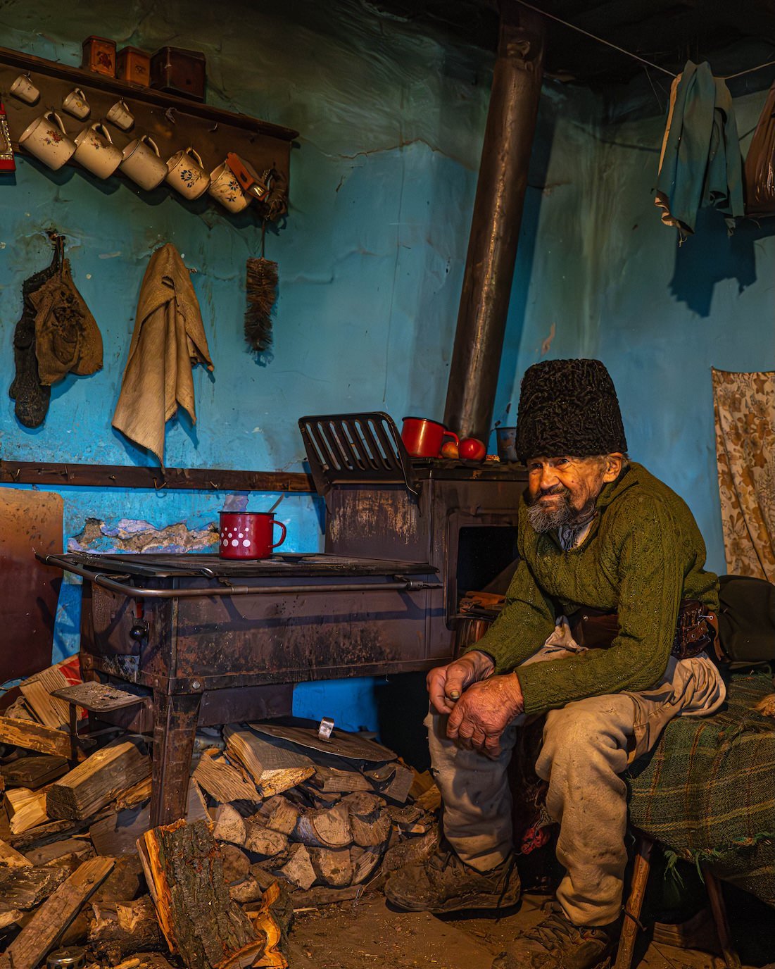 An elderly man with a gray beard and dark hat sitting beside a wood stove in a rustic kitchen, surrounded by stacked firewood, hanging cloth, and decorated with traditional items.