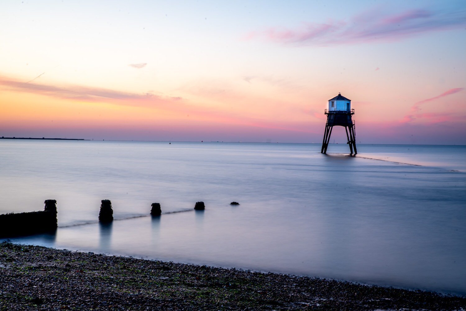 A lighthouse standing in shallow water near the beach at sunset, with colorful sky and calm sea.