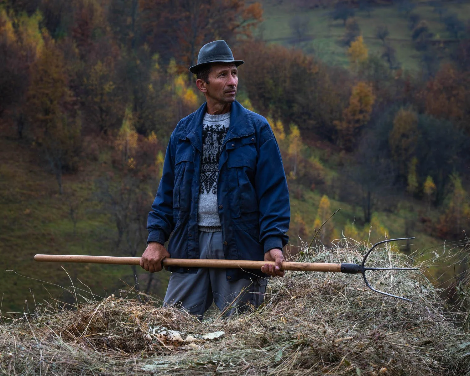 A man outdoors in fall foliage, holding a rake with dry grass and debris, wearing a hat, blue jacket, and sweater, looking to the side.