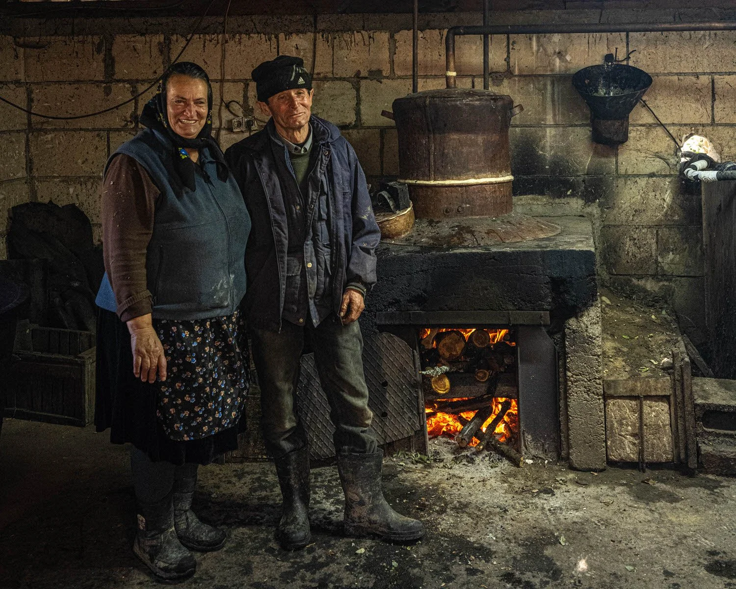 Two people standing in front of a rustic fireplace with a fire burning inside, inside a stone-walled room. The woman on the left is smiling, wearing a headscarf, jacket, and apron, while the man on the right has a neutral expression, wearing a hat, j